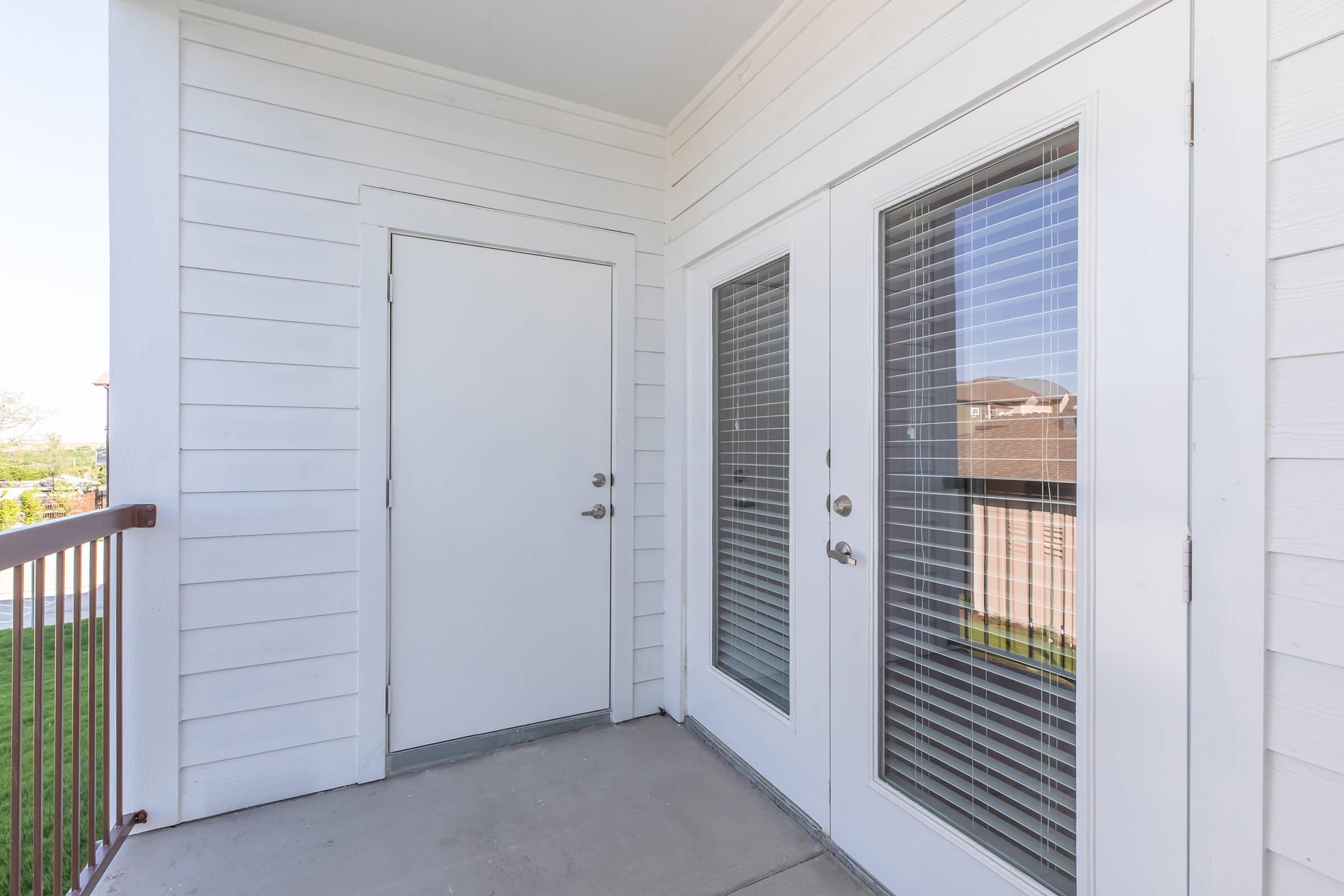 An exterior view of a modern balcony or porch area featuring a plain white door and two large glass doors with blinds. The walls are painted white, and there is a railing visible. The scene suggests a bright, open space with greenery in the background.