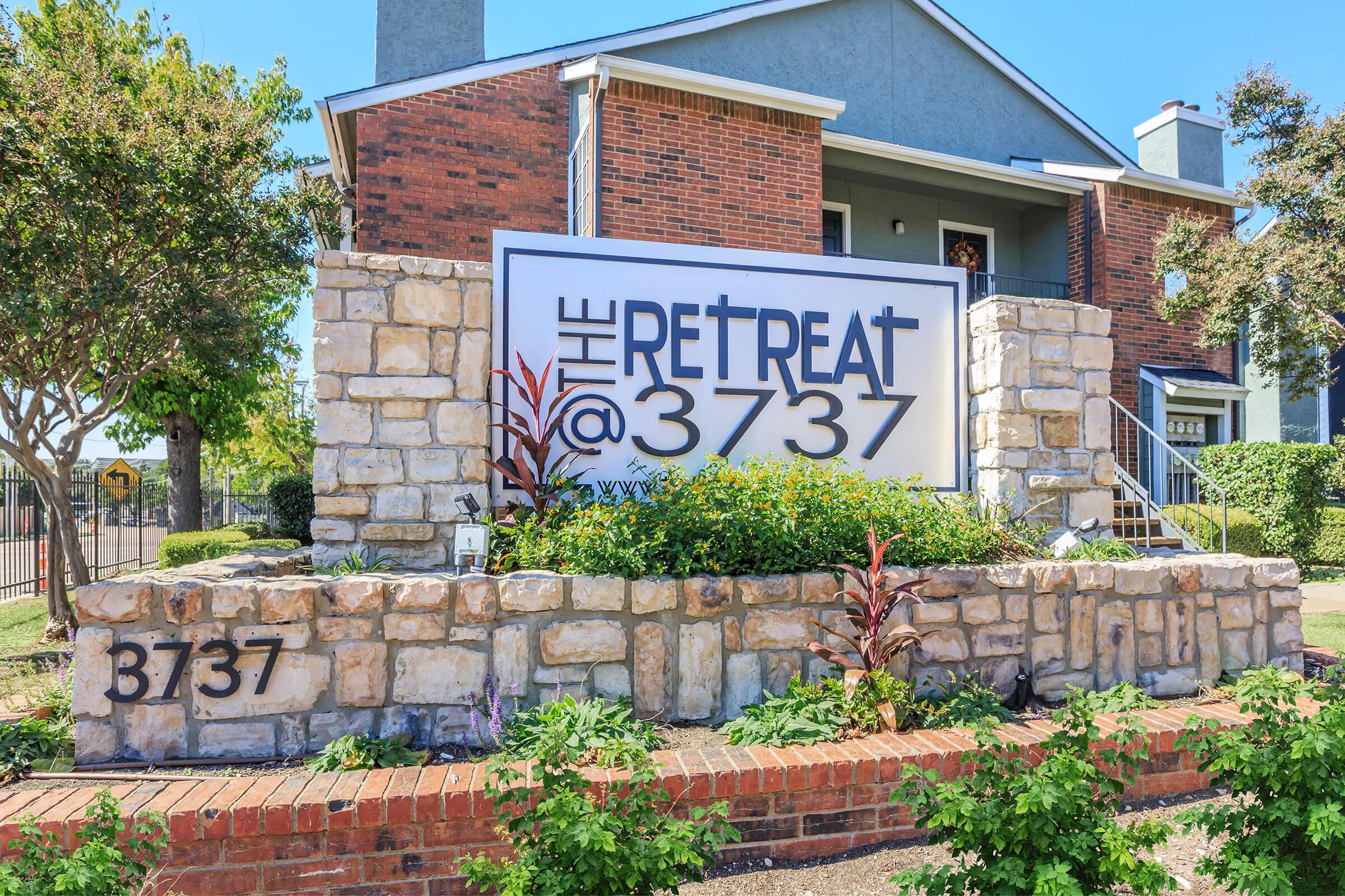 Sign for "The Retreat @ 3737," featuring bold lettering on a large white background, set against a stone and red brick facade. The entrance is landscaped with green plants and colorful flowers, and there are steps leading up to the building in a sunny outdoor setting.