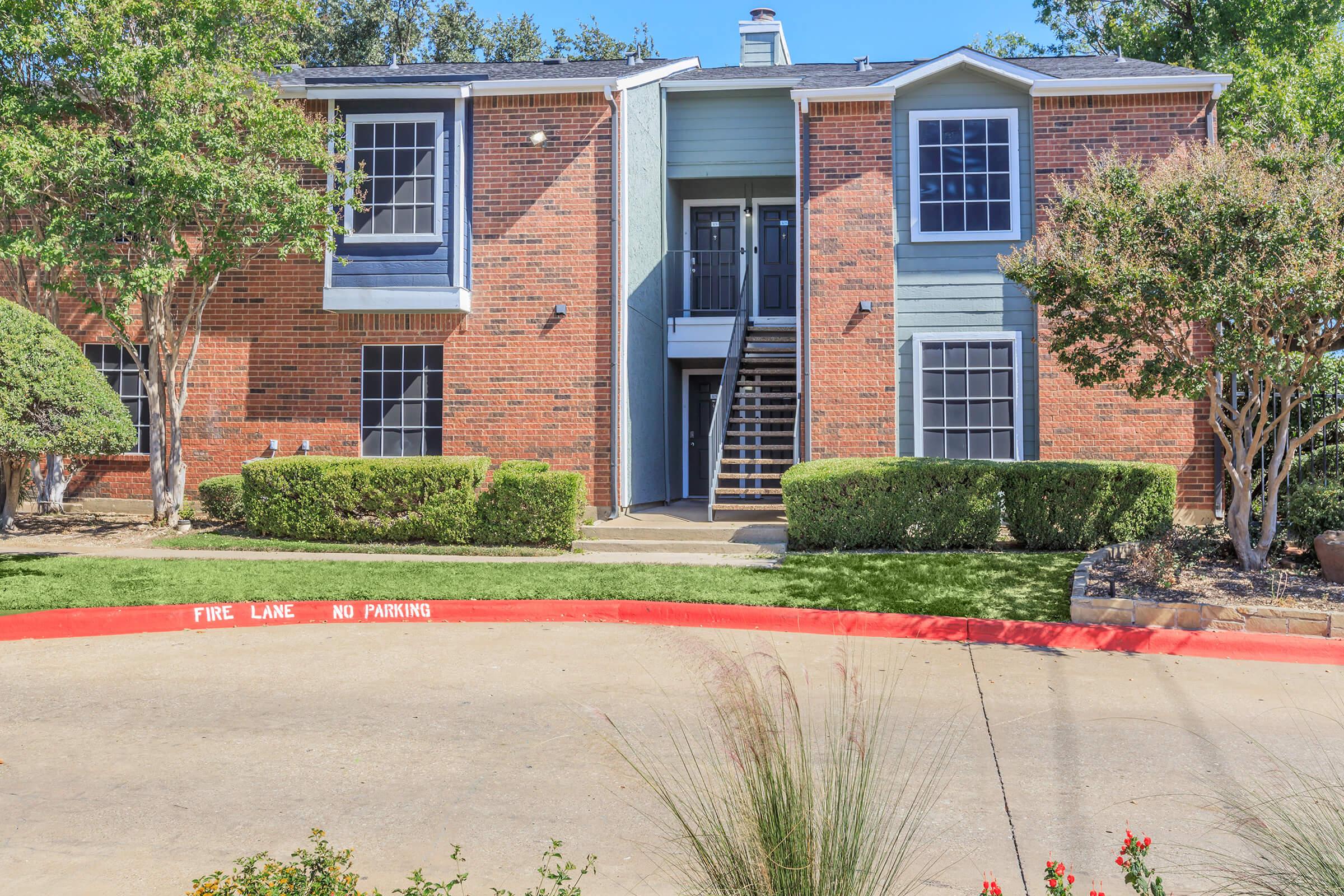 A brick apartment building with blue accents, featuring two upper-level windows and a central staircase. The front yard has manicured shrubs and a circular driveway with a painted warning for "FIRE LANE NO PARKING." Trees line the property, providing shade.