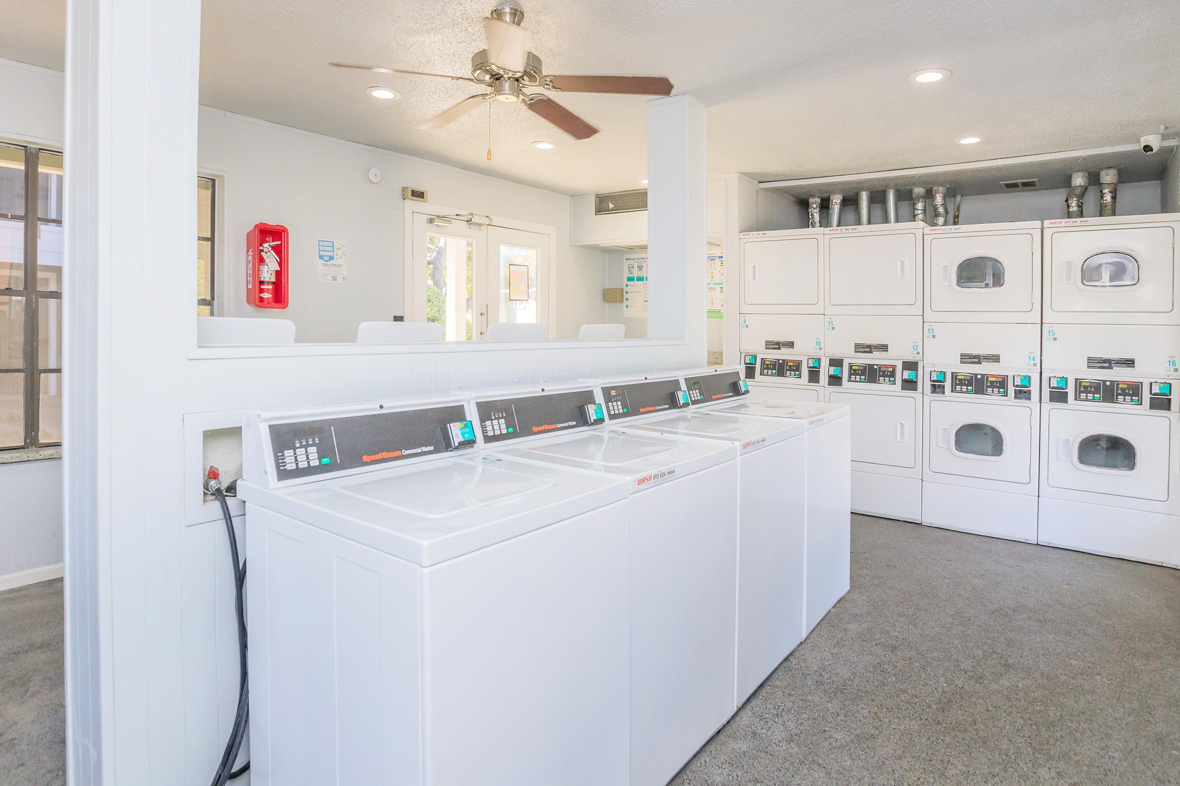 A clean laundry room featuring several white washing machines and dryers, with a ceiling fan above. The room has light-colored walls, windows allowing natural light, and a fire extinguisher mounted on the wall. It creates a well-lit and inviting atmosphere for laundry tasks.