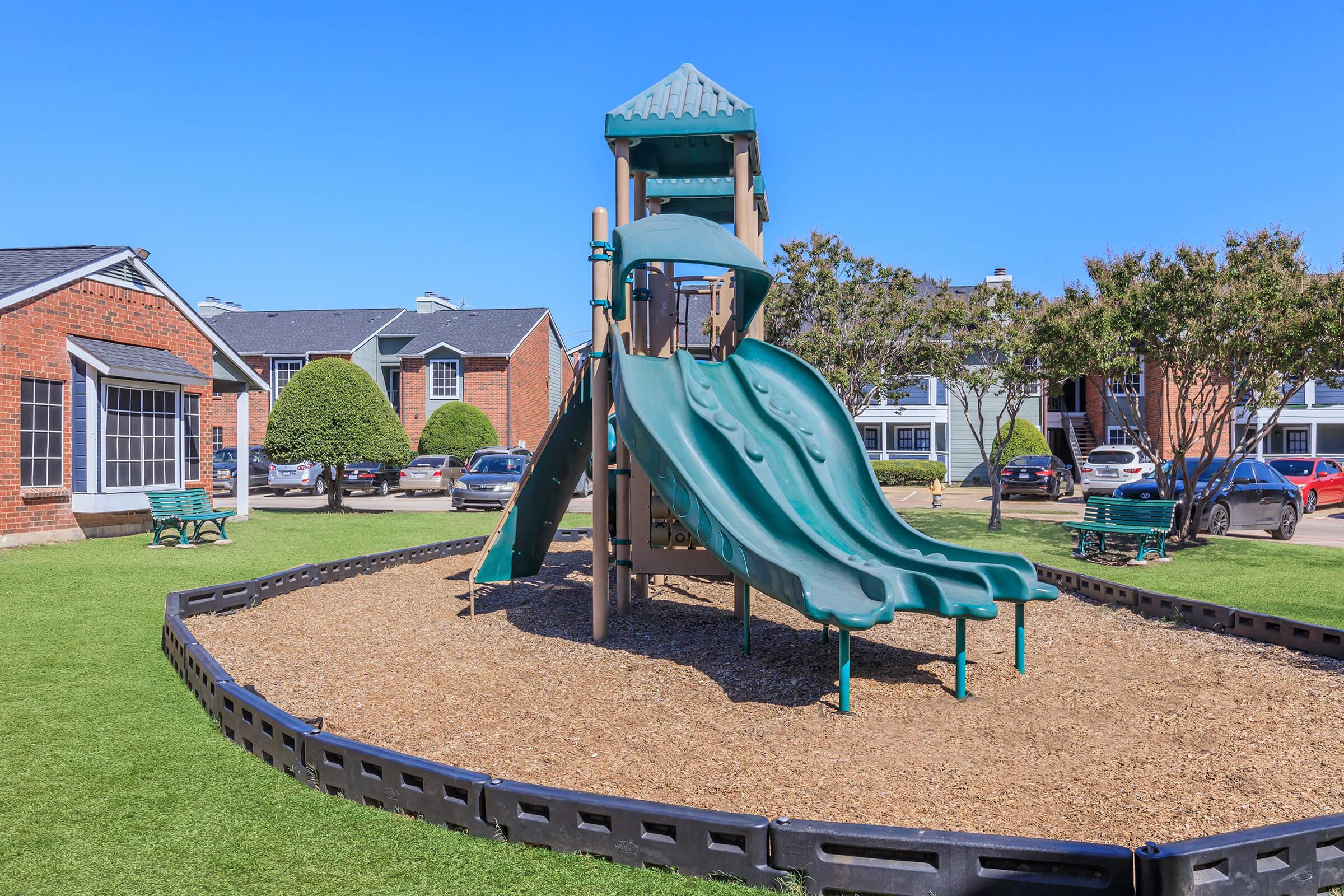 A playground featuring a large green slide structure on a bed of mulch, surrounded by a grassy area. Nearby, there are benches and parked cars, with apartment buildings visible in the background under a clear blue sky.