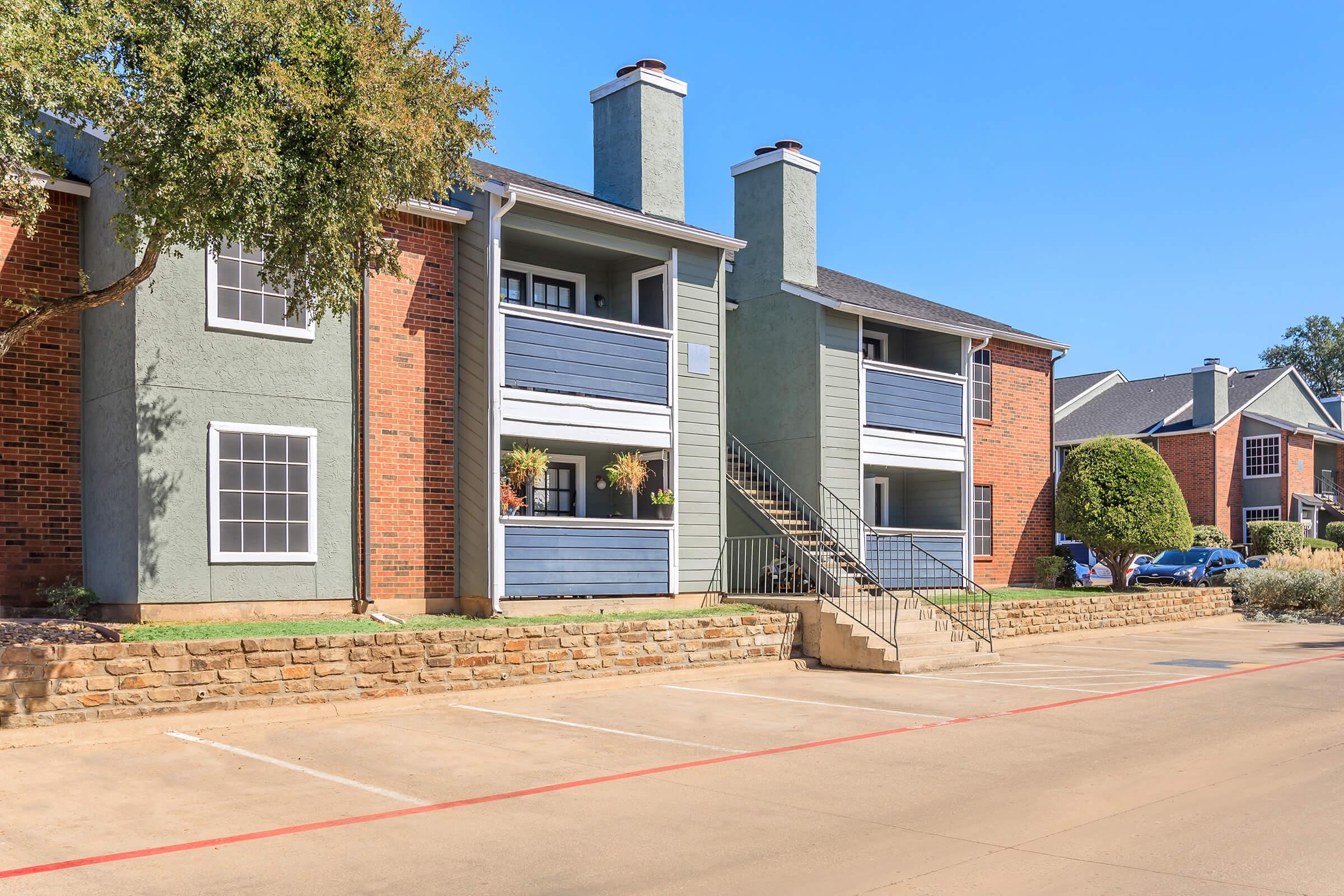Exterior view of a multi-unit apartment building featuring a mix of brick and painted siding. The building has two levels with balconies adorned with plants. Stairs lead to the upper level. A paved parking area is in front, with greenery in the surrounding landscape and a clear blue sky overhead.