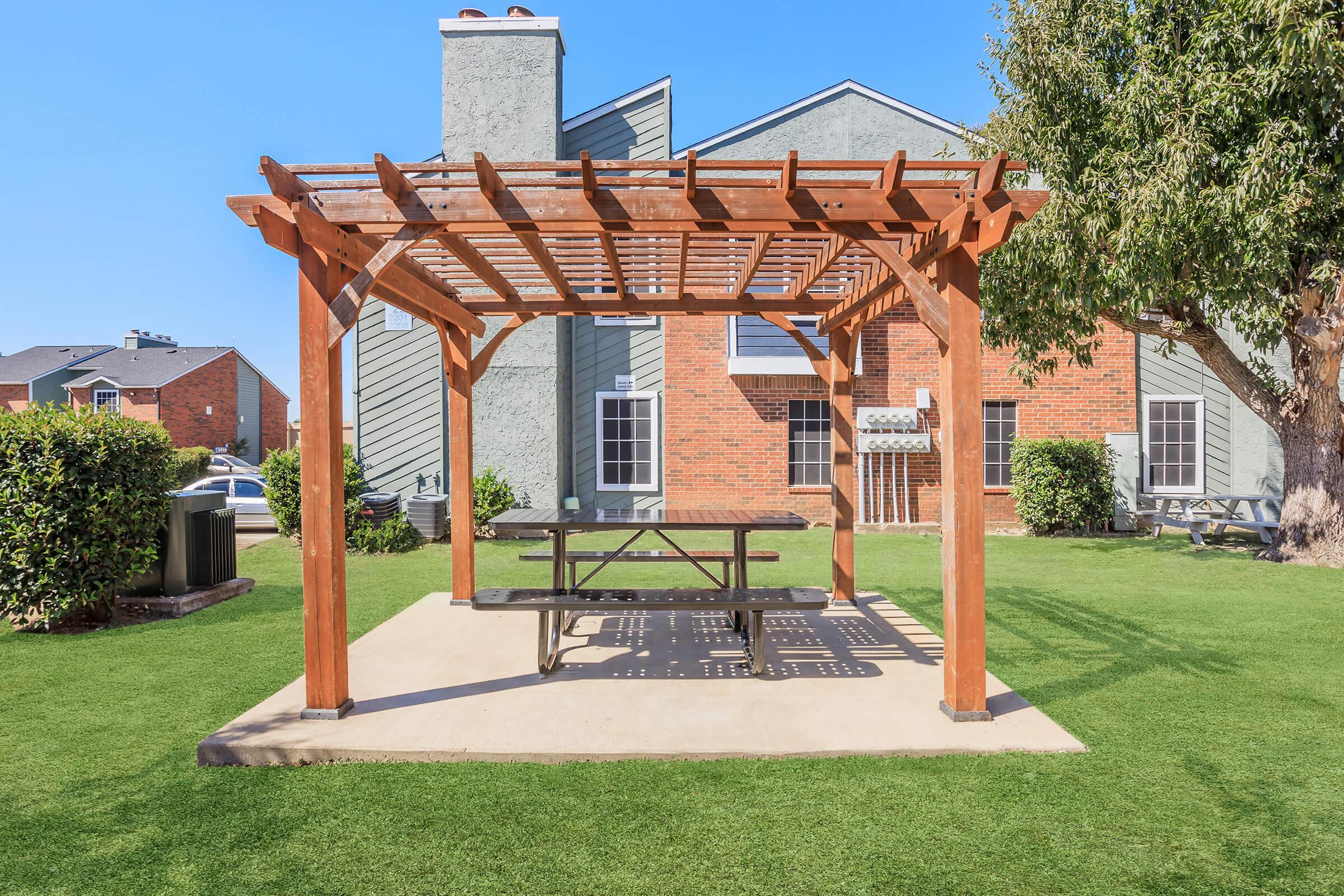 A wooden pergola with a picnic table underneath, situated on a grassy area. In the background, there are two residential buildings with brick and gray exteriors. The sky is clear and blue, creating a bright and inviting atmosphere.