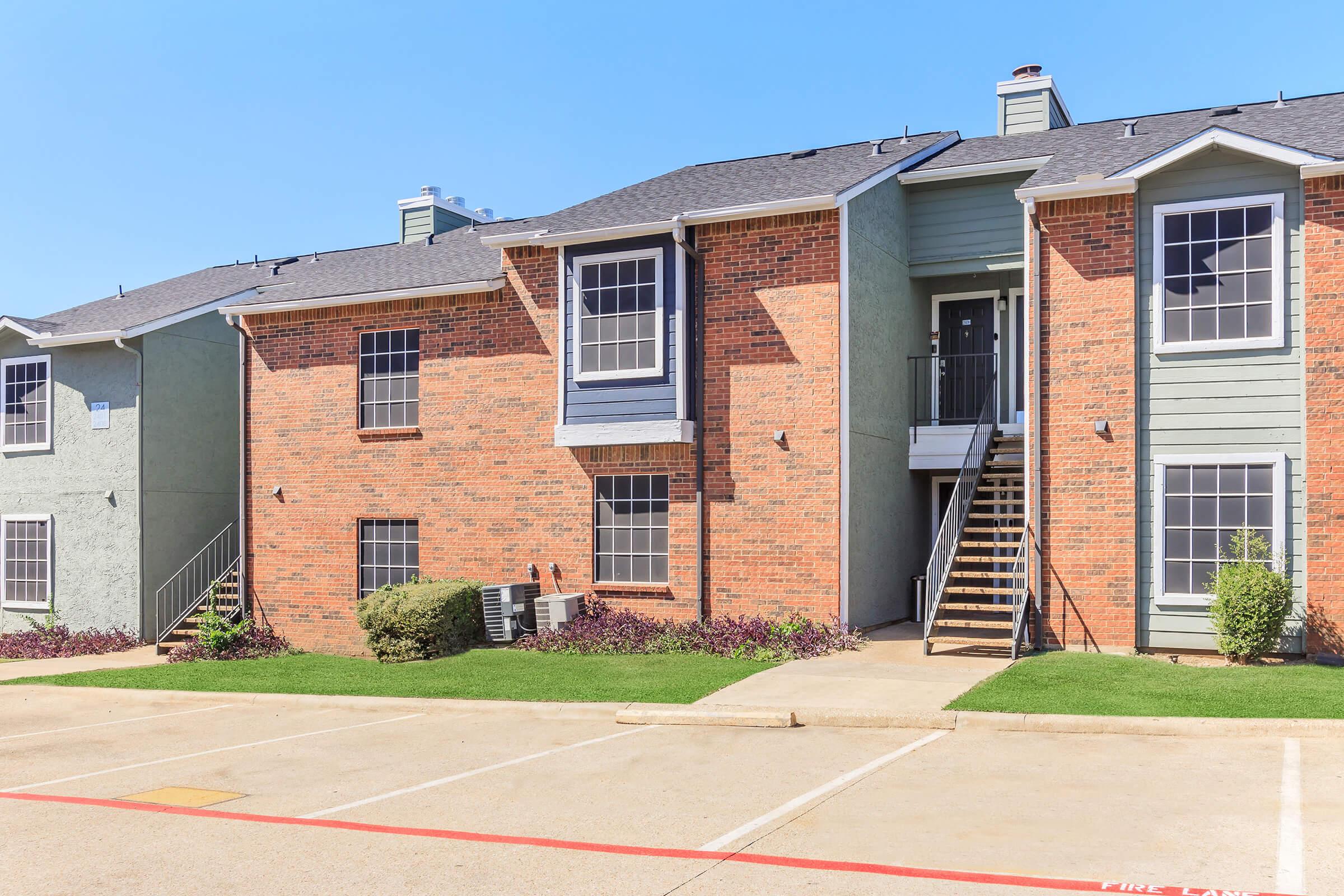 Two-story apartment building featuring a combination of brick and green siding. The facade includes large windows and a staircase leading to the upper level. A landscaped area with small shrubs is visible in front of the building, along with a paved parking space. The sky is clear and blue.