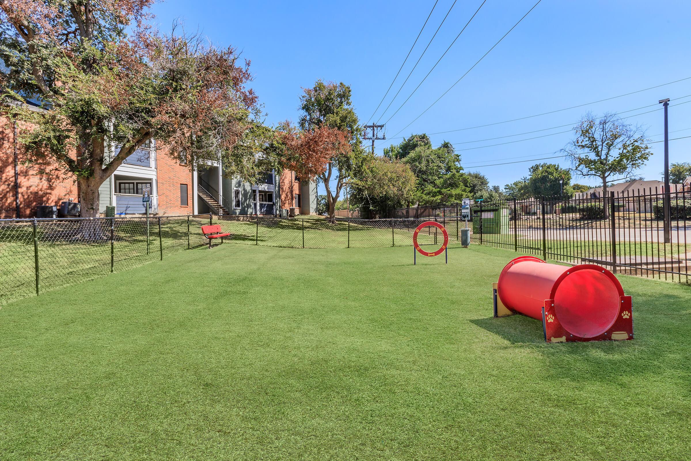 A dog park featuring a grassy area with a red agility tunnel and a hoop. A bench is positioned nearby, and there are trees in the background. The park is enclosed by a fence, with utility poles and buildings visible in the distance under a clear blue sky.