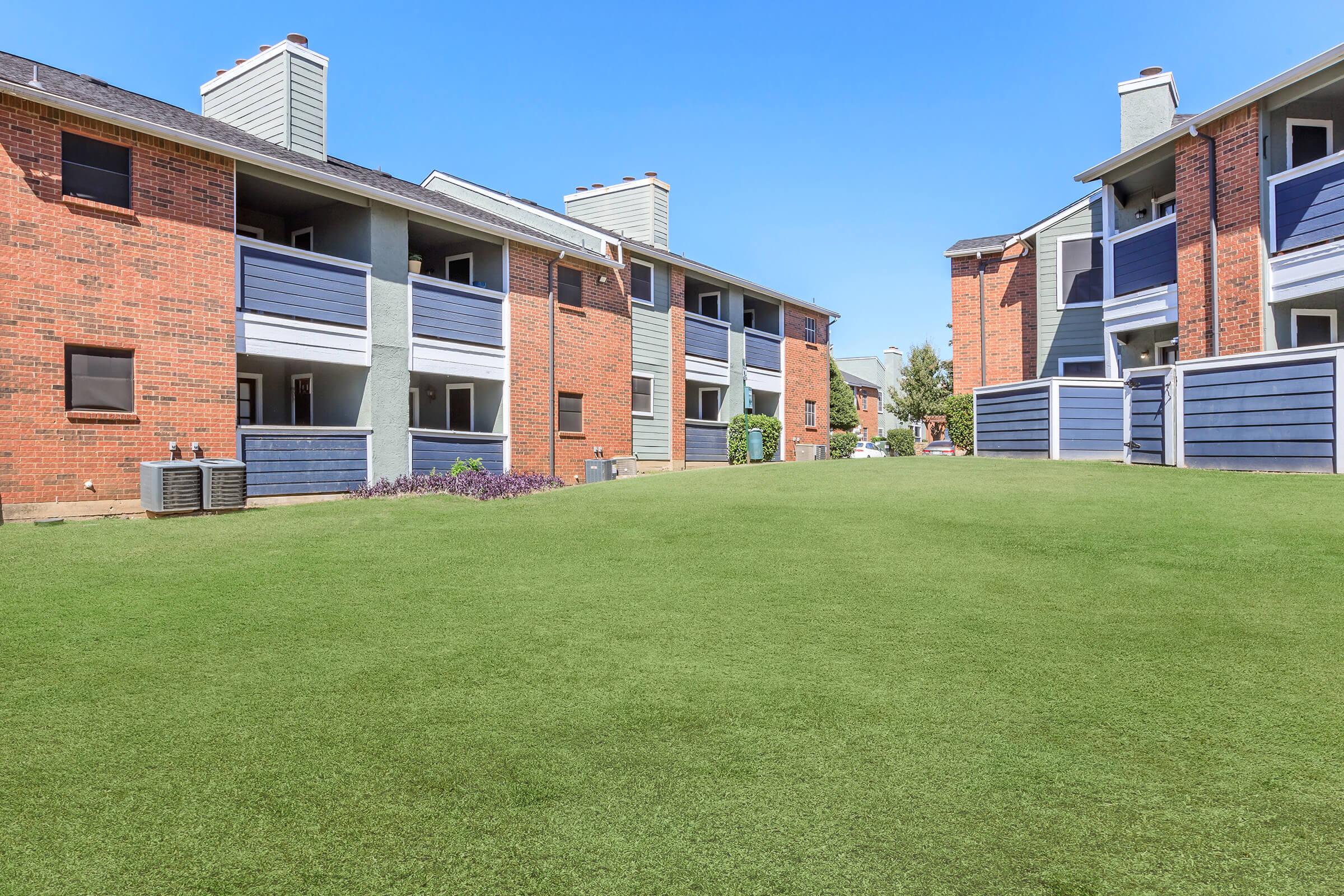 View of a well-maintained apartment complex featuring two buildings with a mix of brick and blue siding. The grassy lawn in the foreground is neatly trimmed, and the overall setting is sunny and inviting. Air conditioning units are visible on the left side of the image.