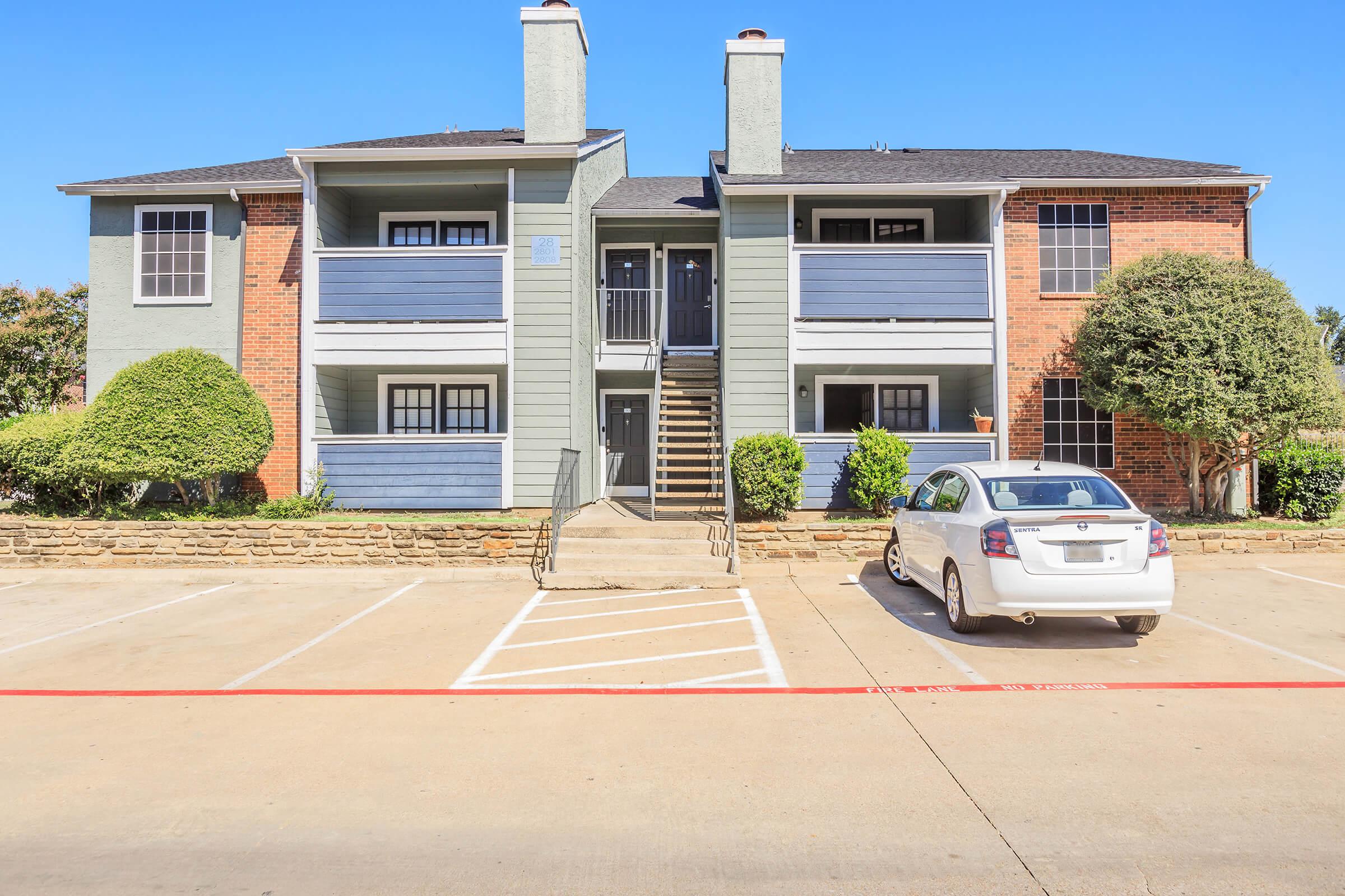 A two-story apartment building with a combination of brick and gray siding. There are balconies on the upper level and a set of stairs leading to the entrance. The parking lot in front has a white car parked next to the building, with neatly trimmed bushes and a clear sky in the background.