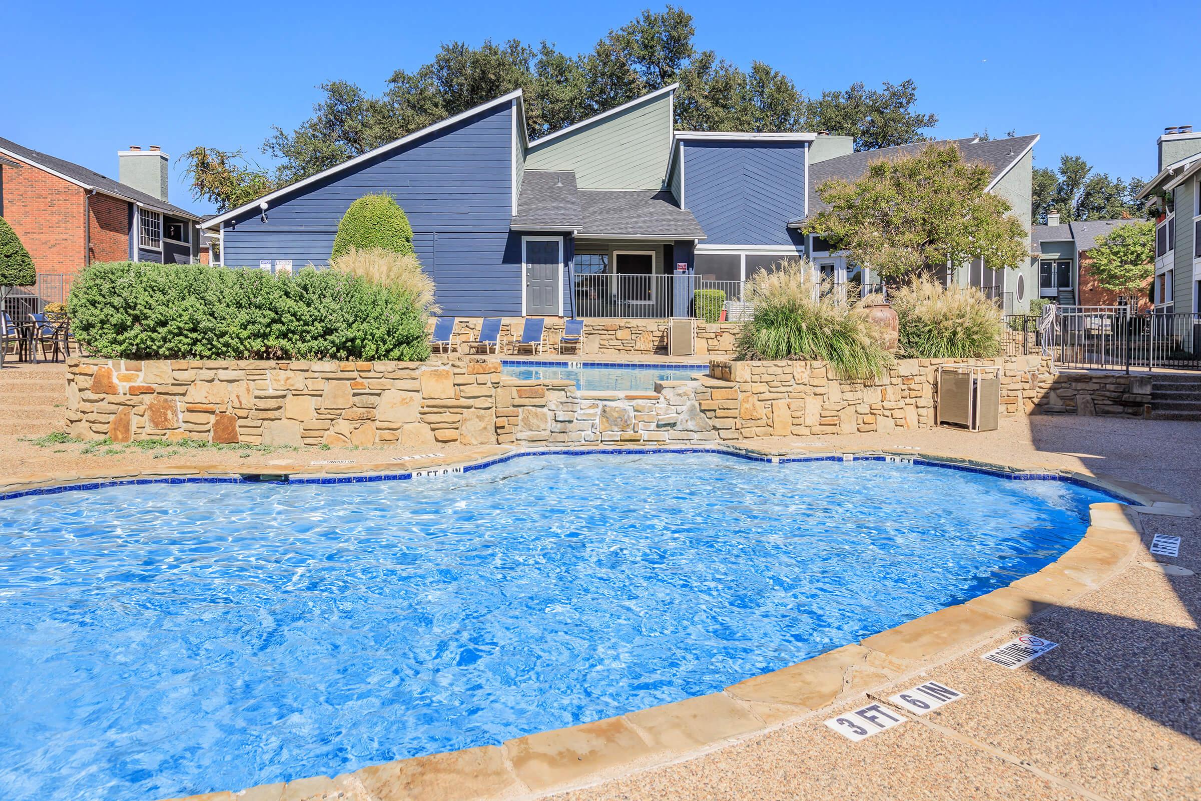 A sparkling blue swimming pool surrounded by a stone patio and lounge chairs. In the background, there are modern buildings with a mix of gray and blue exteriors, along with well-maintained landscaping featuring shrubs and trees. The area appears sunny and inviting, perfect for relaxation.