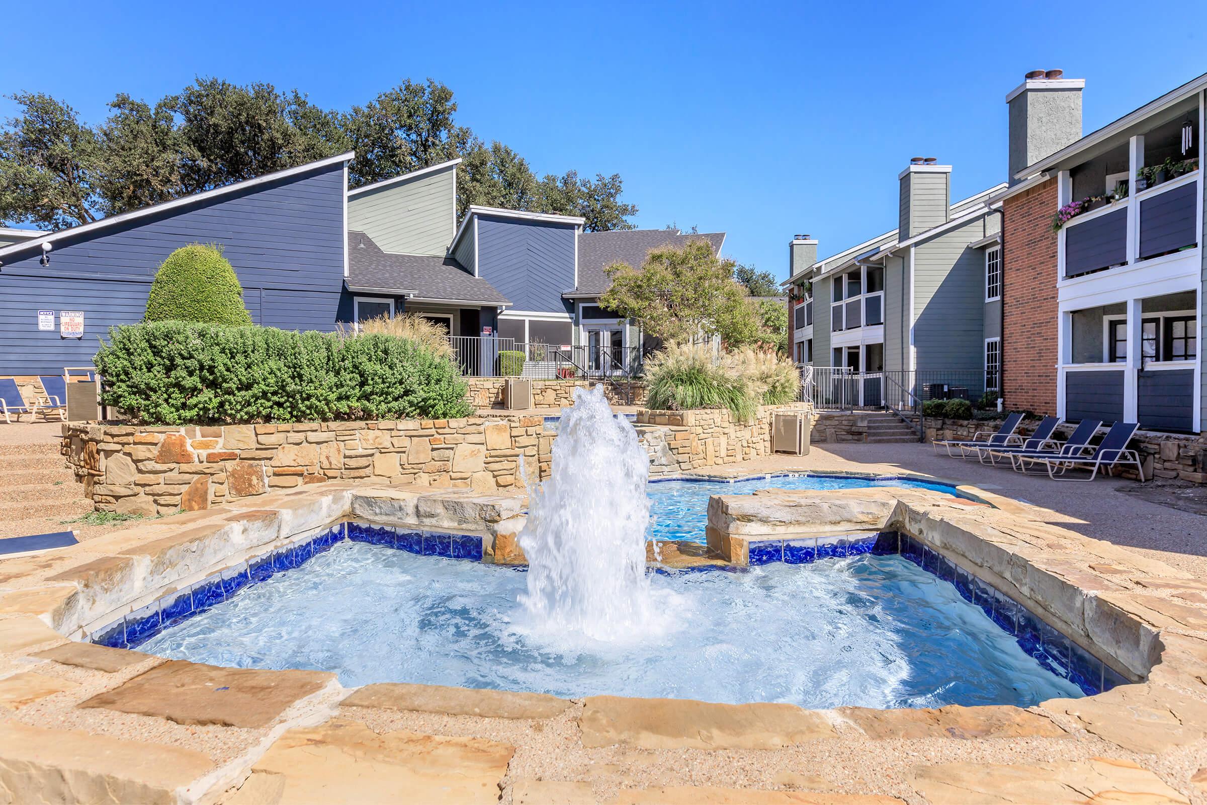 A clear blue pool area featuring a stone fountain in the center, surrounded by lush greenery and well-maintained landscaping. Nearby, there are lounge chairs for relaxation, and apartment buildings are visible in the background under a clear blue sky.