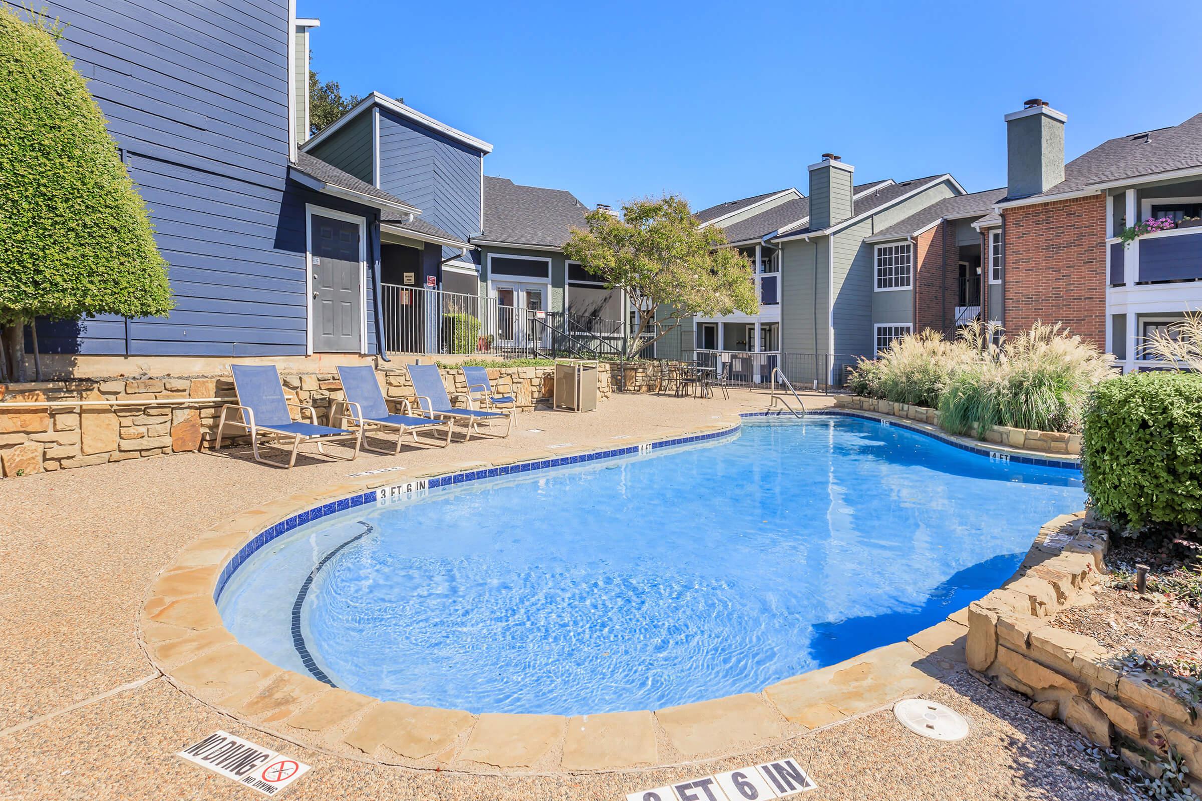 A well-maintained outdoor swimming pool surrounded by lounge chairs, with a vibrant landscape of shrubs and grass. The setting features modern apartment buildings in the background under a clear blue sky. The pool has a gentle curve and a depth marker indicating a maximum depth of 6 feet.