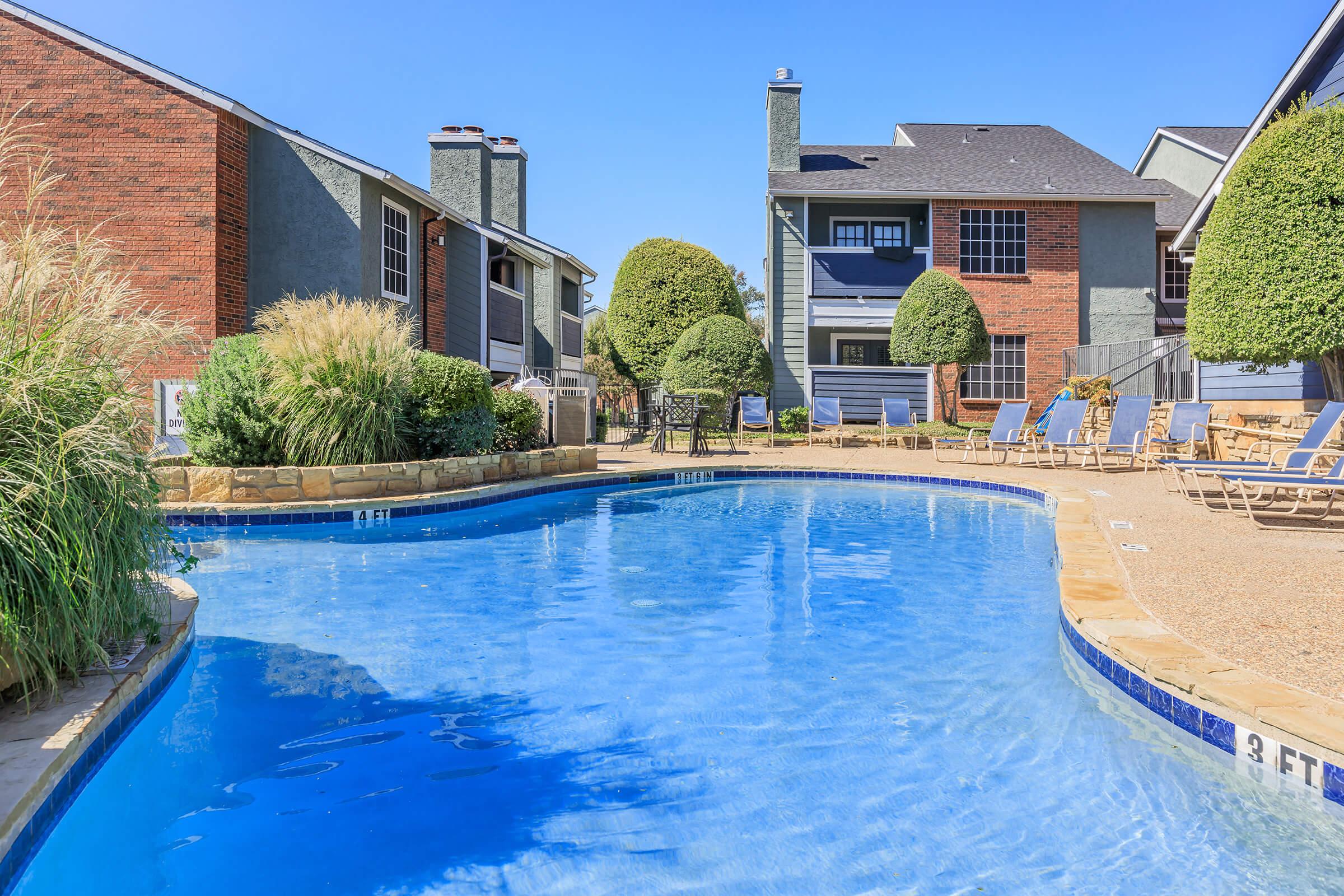 A clear swimming pool surrounded by landscaped greenery and lounge chairs. In the background, residential buildings with a mix of brick and siding are visible under a clear blue sky. The pool area features a shallow end marked with a depth indicator.