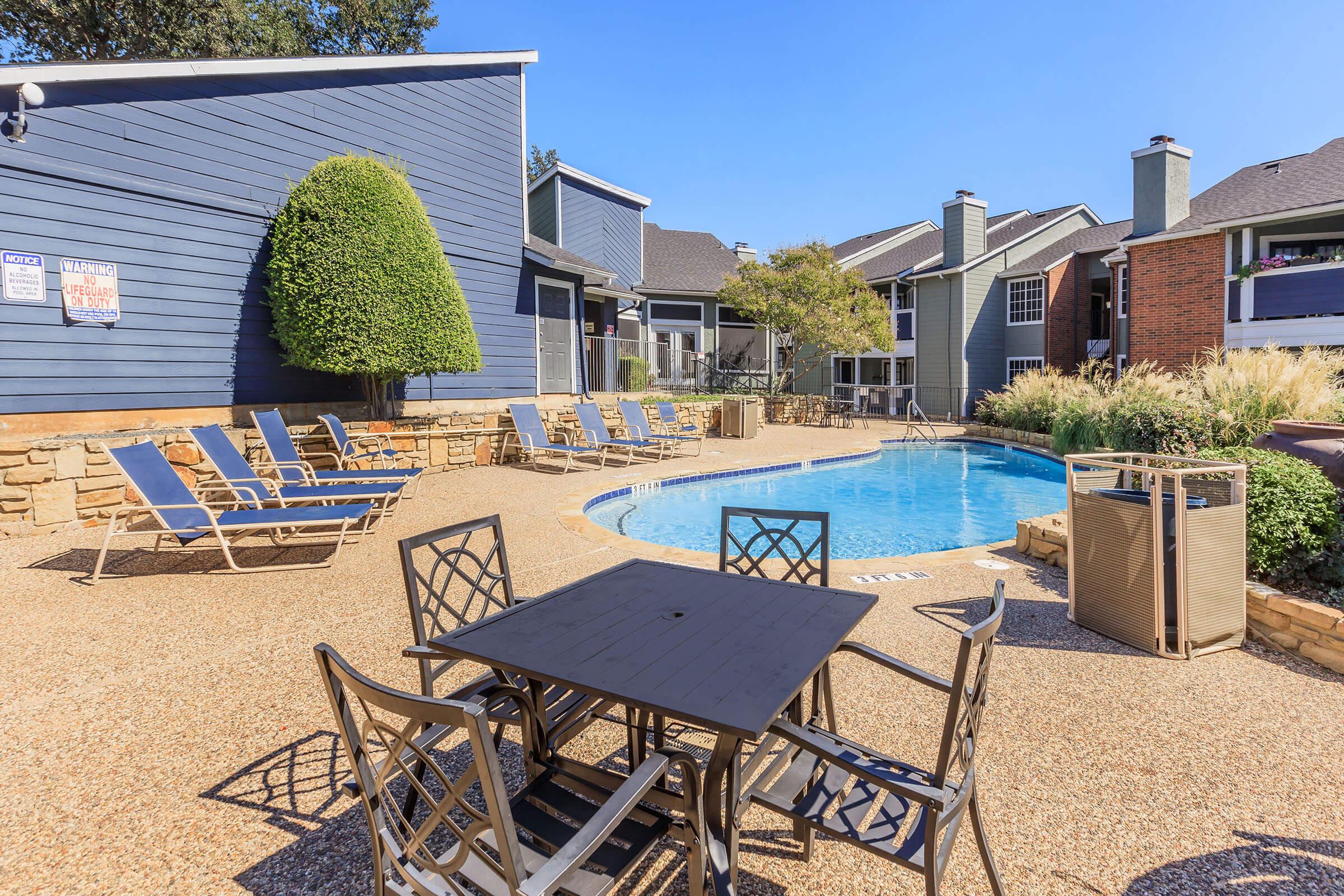 A swimming pool surrounded by lounge chairs and a table with chairs, set in a landscaped area with buildings in the background. The scene is under a clear blue sky, creating a relaxing outdoor atmosphere.