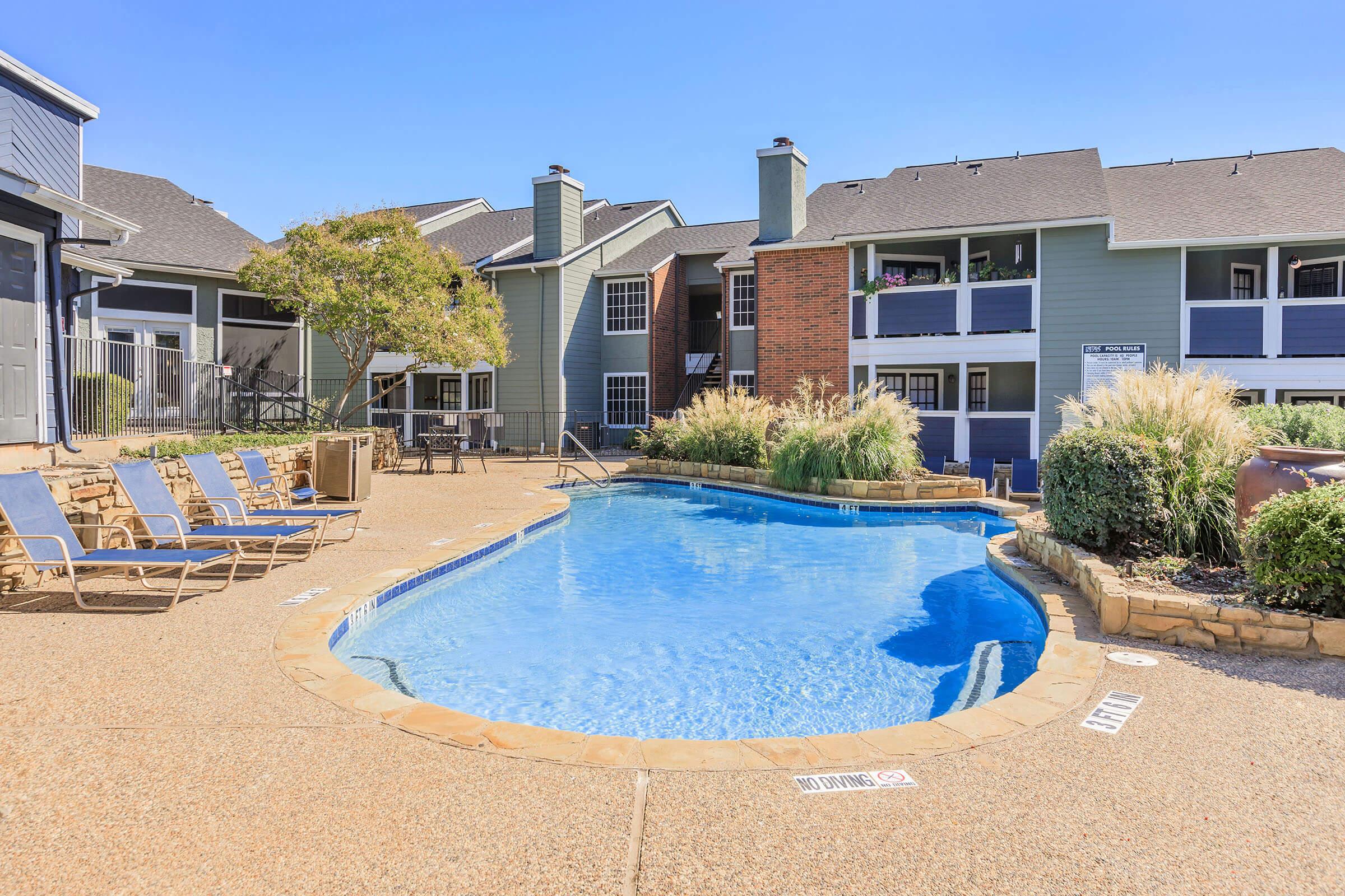 A sunlit swimming pool surrounded by lounge chairs and landscaped greenery, with apartment buildings in the background. The pool has a curved shape and is set in a residential area, creating a relaxing atmosphere for residents.
