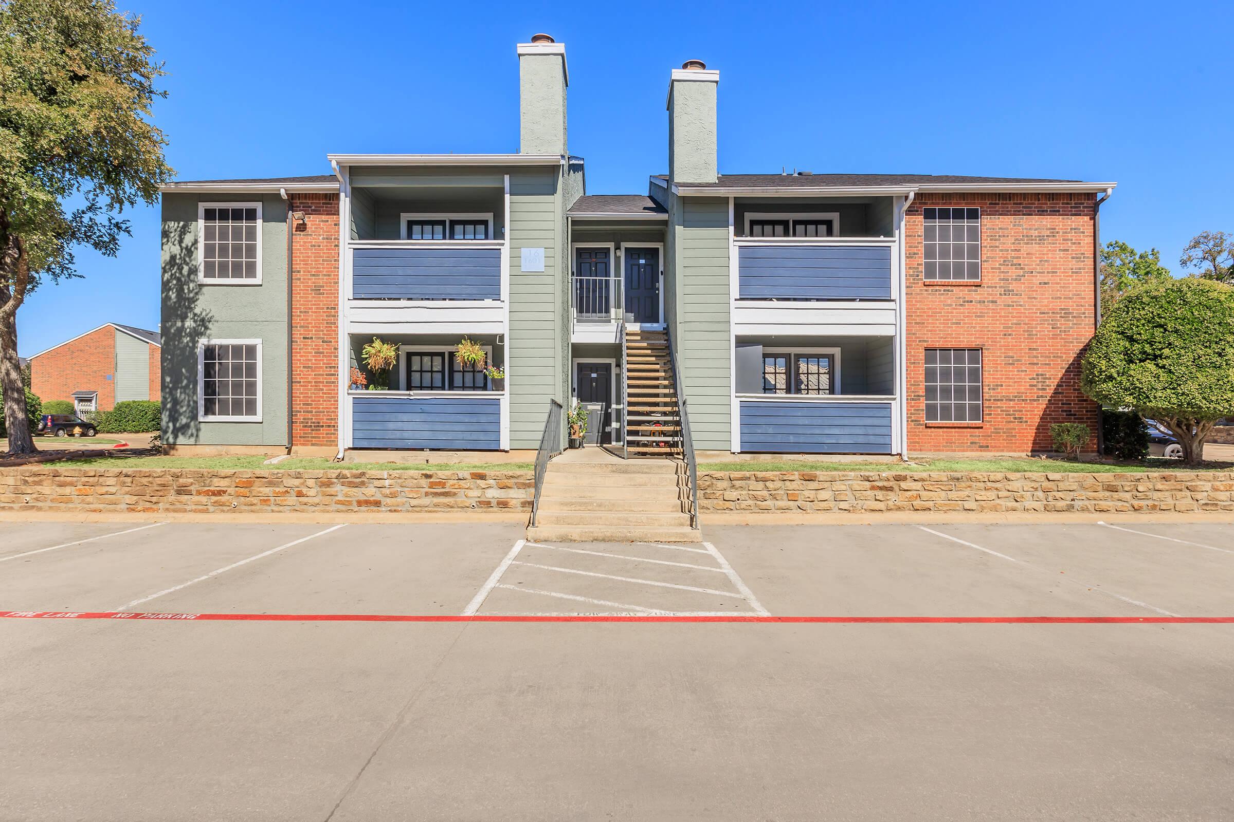 A two-story apartment building with a mix of brick and blue siding. The front porch has stairs leading up to the second floor, with planters on either side. There are several parking spots in front, and the sky is clear and blue, indicating a sunny day.