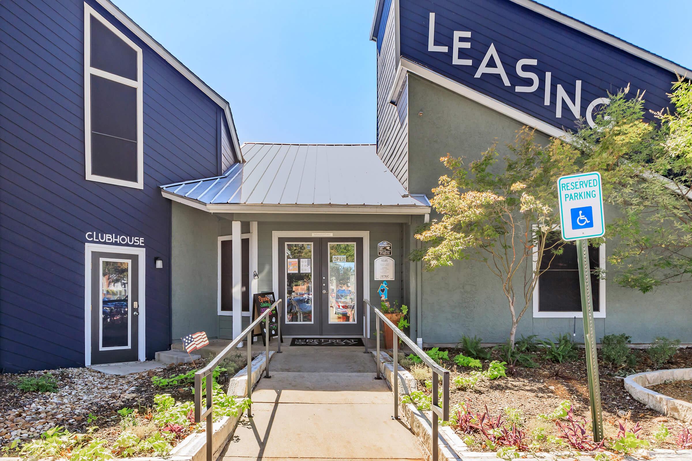 A modern leasing office with a blue and grey exterior. The entrance features a ramp, signage reading "LEASING" and "CLUBHOUSE," and manicured landscaping with plants. A reserved parking sign is visible nearby. The building has large windows that provide a welcoming atmosphere.