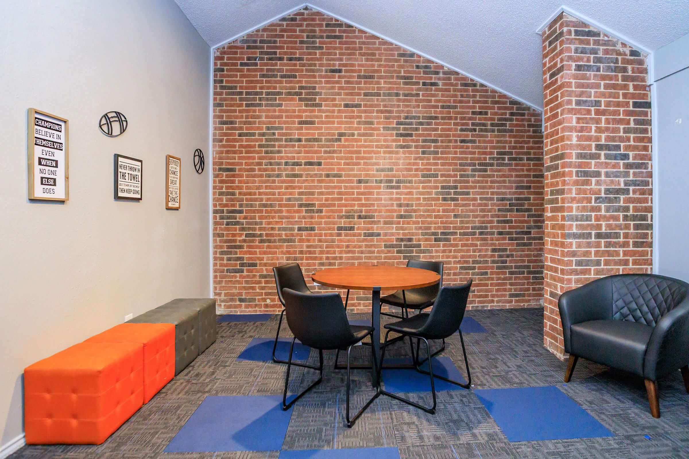 A modern meeting space featuring a round wooden table surrounded by black chairs. The floor has blue and gray tiles, and one side of the wall is adorned with a textured brick pattern. There are two decorative pieces on the wall, adding a stylish touch to the room.
