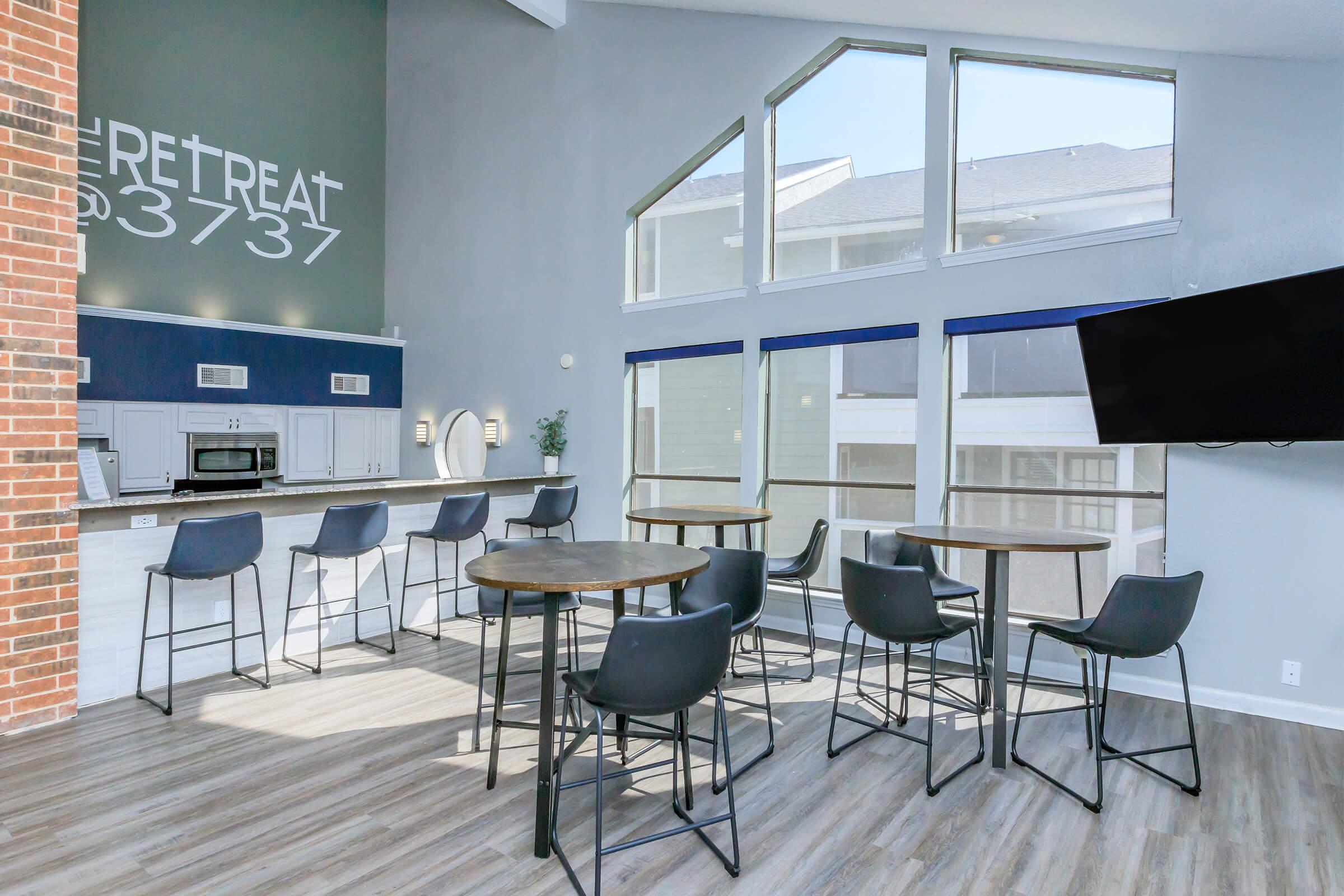 A modern lounge area featuring a bar with high stools, a kitchenette, and large windows allowing natural light. The walls are painted in light colors, and there is a television mounted on one wall. The space has a welcoming and contemporary atmosphere suitable for social gatherings.