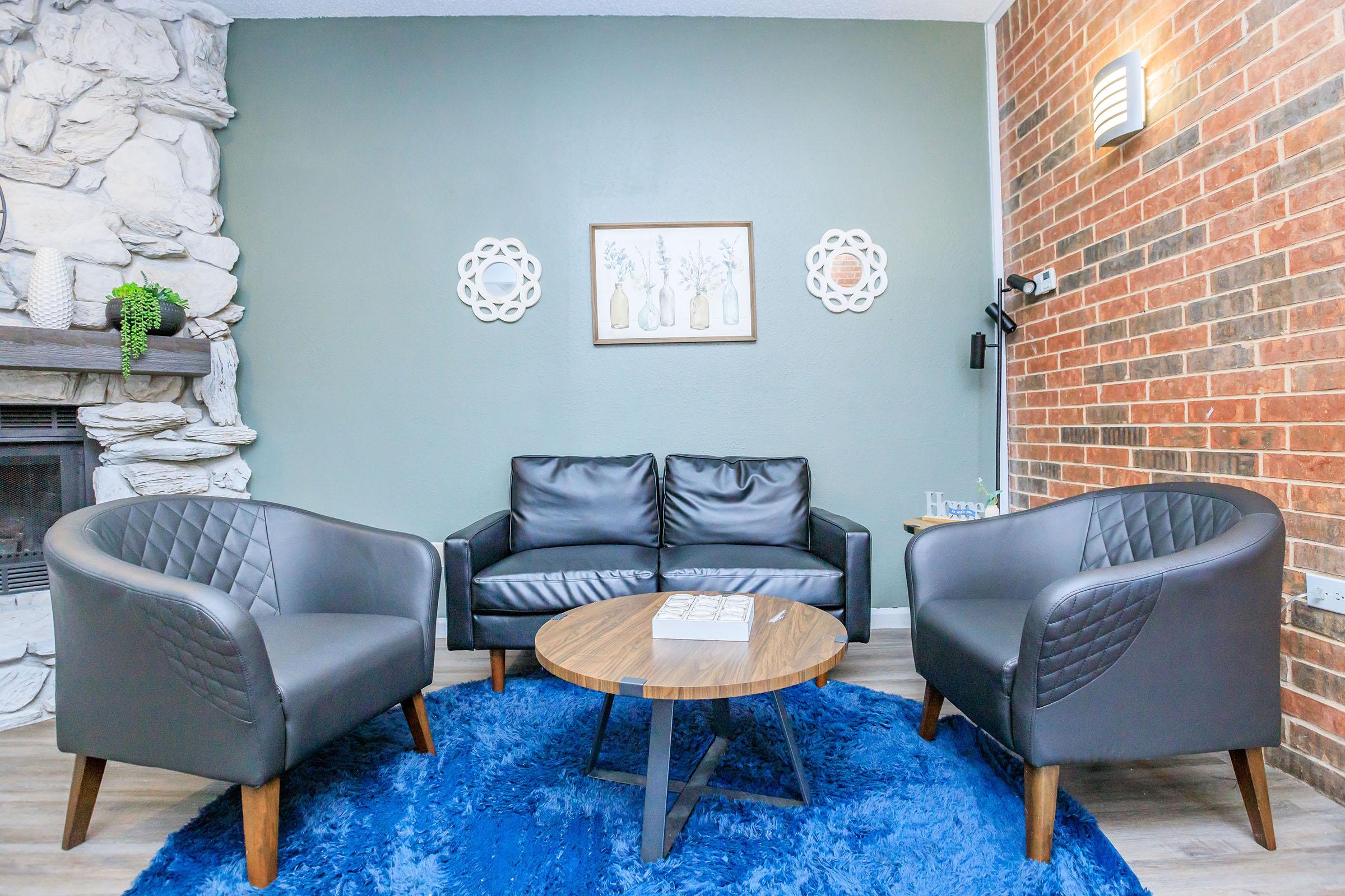 A cozy living room featuring a black leather sofa and two matching armchairs with diamond-patterned backs. In the center, there is a round wooden coffee table on a blue area rug. The walls have a mix of stone and brick accents, and framed artwork with floral motifs is displayed above the sofa.