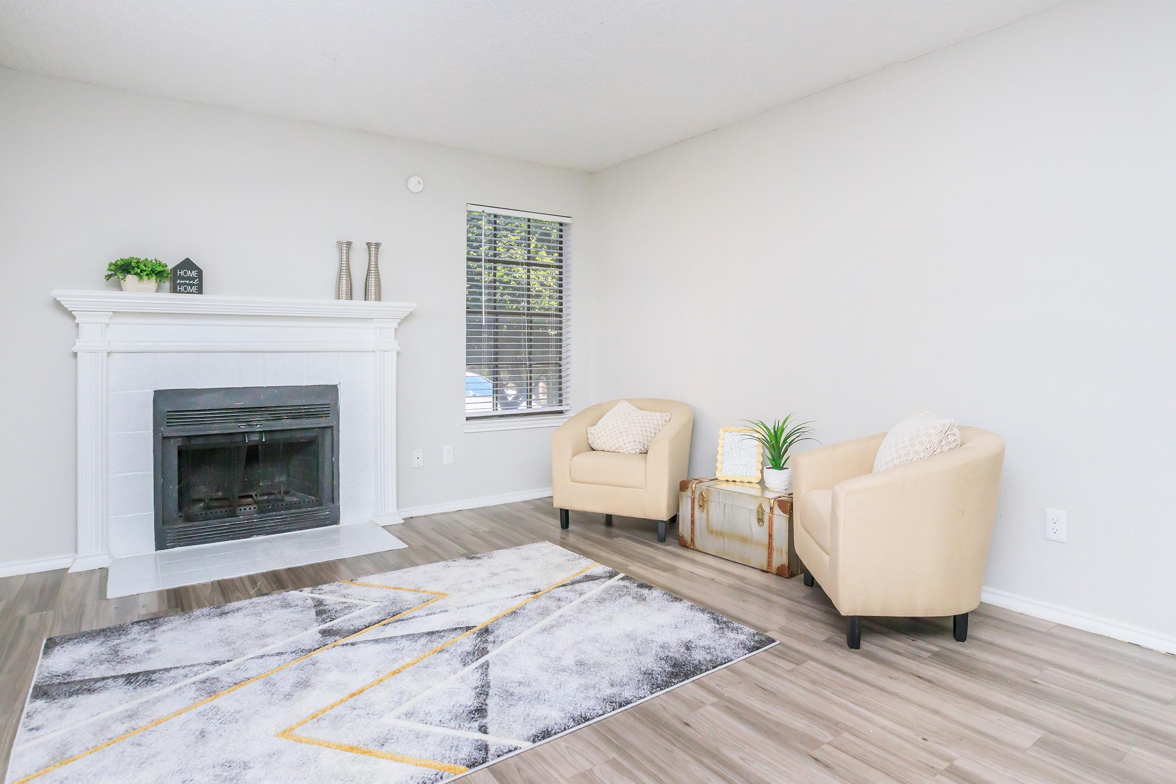 A cozy living room featuring a white fireplace, two beige armchairs, and a decorative area rug with geometric patterns. A small table and a plant add a touch of greenery. Large window with blinds allows natural light to brighten the space. Walls are painted in a light color, creating an inviting atmosphere.