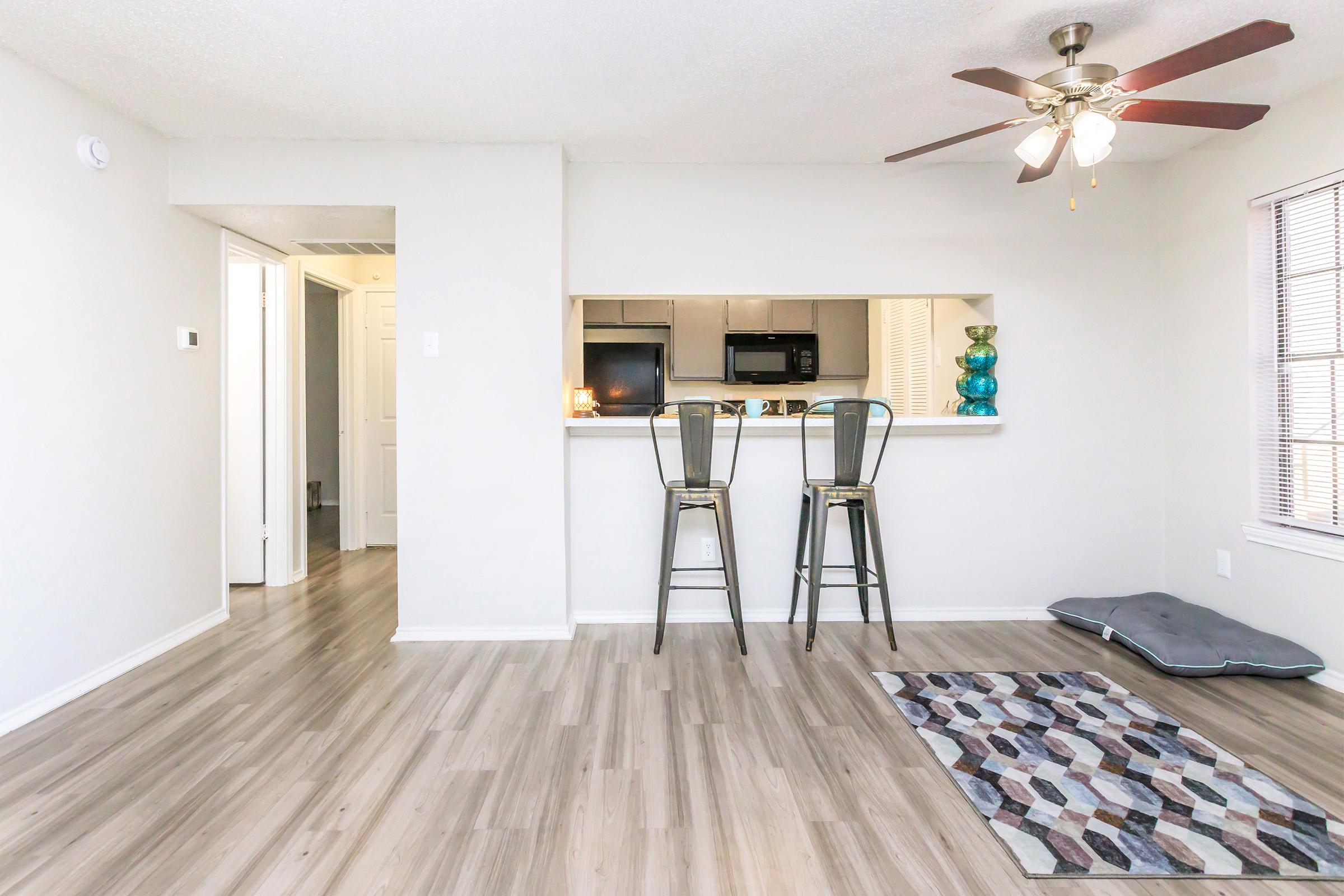 A bright, modern living space featuring a light gray wall, wooden flooring, and a small area rug. Two bar stools are positioned at a kitchen counter with a visible microwave and a blue decorative vase. A fan hangs from the ceiling, and a hallway leads to another room, creating an open and inviting atmosphere.