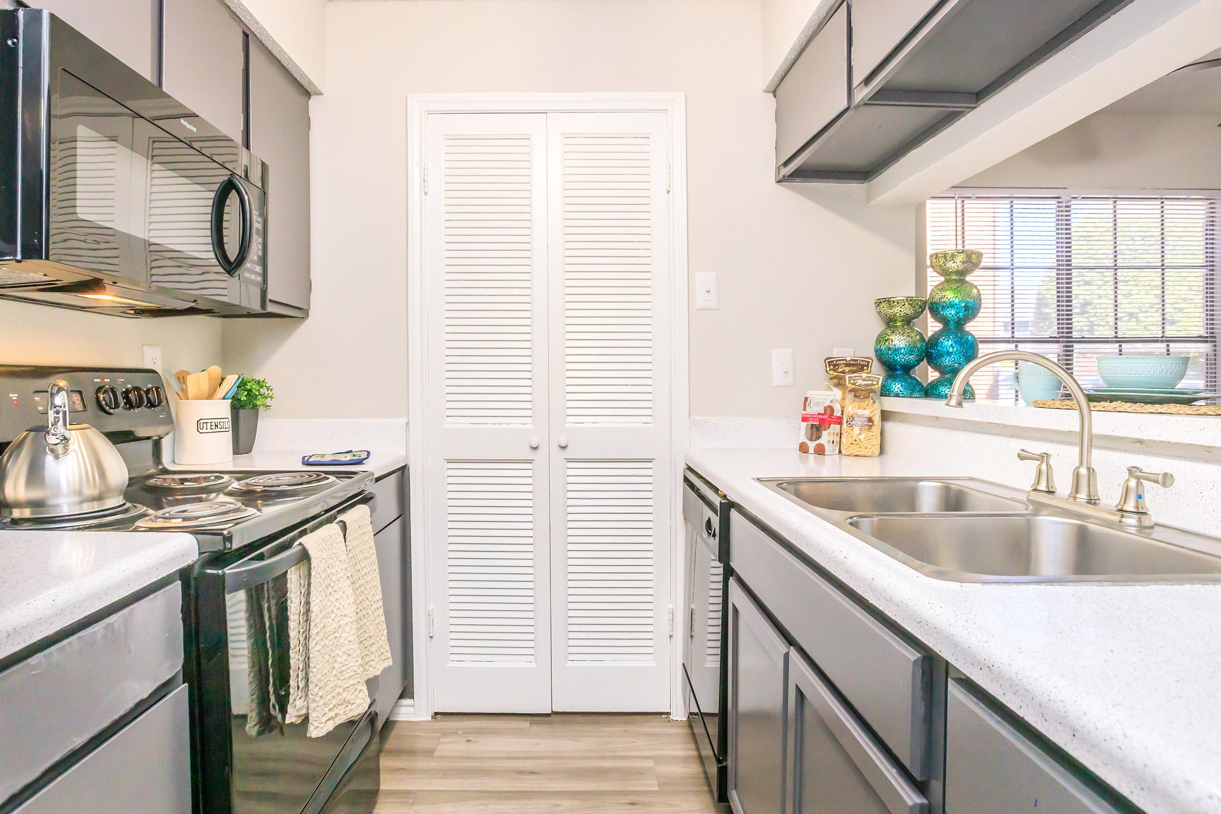 Bright kitchen featuring dark cabinets, stainless steel appliances, and a white countertop. Includes a double sink with a modern faucet, a decorative glass bowl, and a pantry door with louvered panels. Sunlight streams through a window, enhancing the inviting atmosphere.