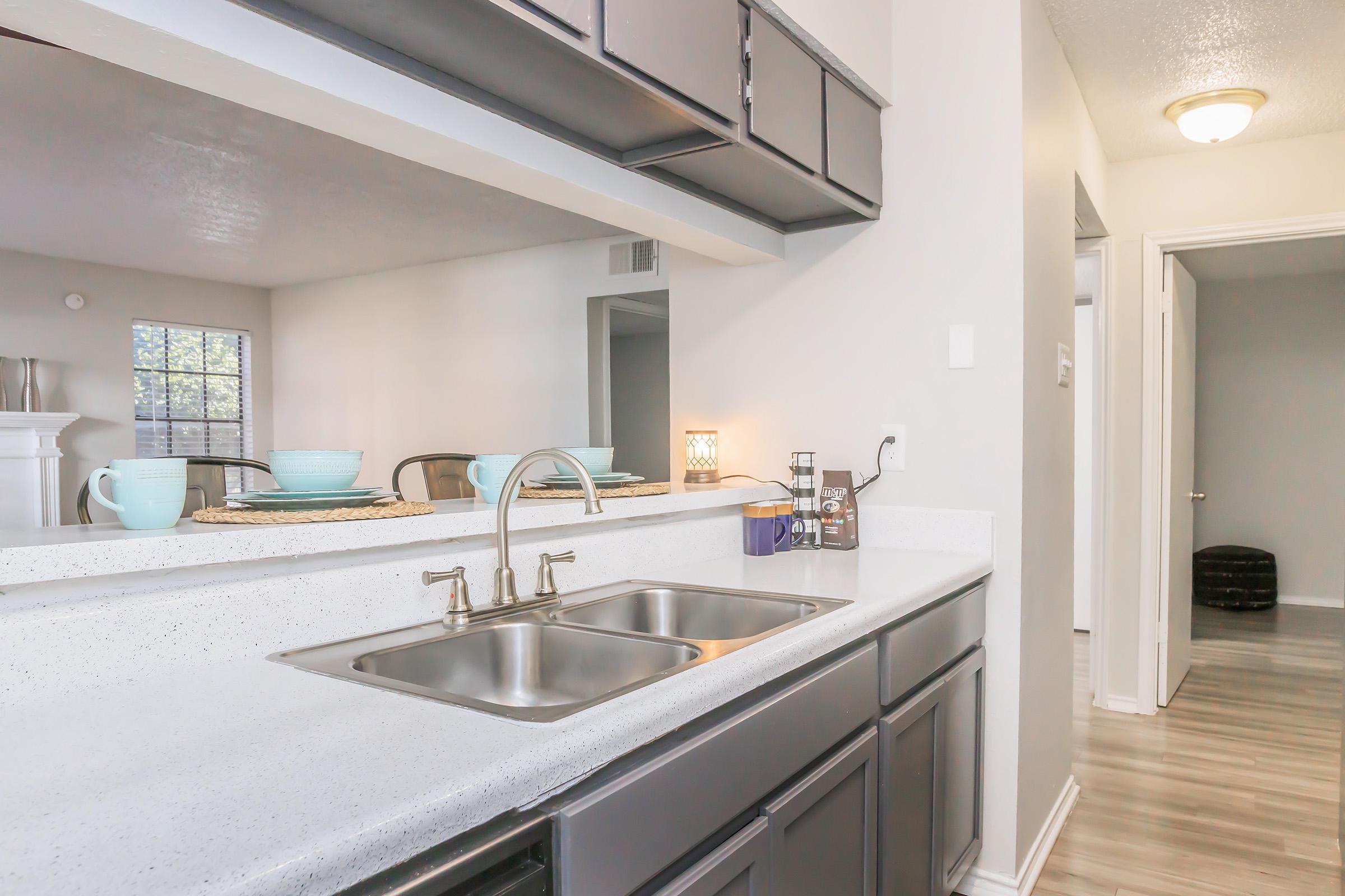 A modern kitchen featuring a double sink, dark cabinets, and a countertop. In the background, there is a dining area with a table set with plates and cups. Natural light enters through a window, and a doorway leads to another room. The overall decor is clean and contemporary.