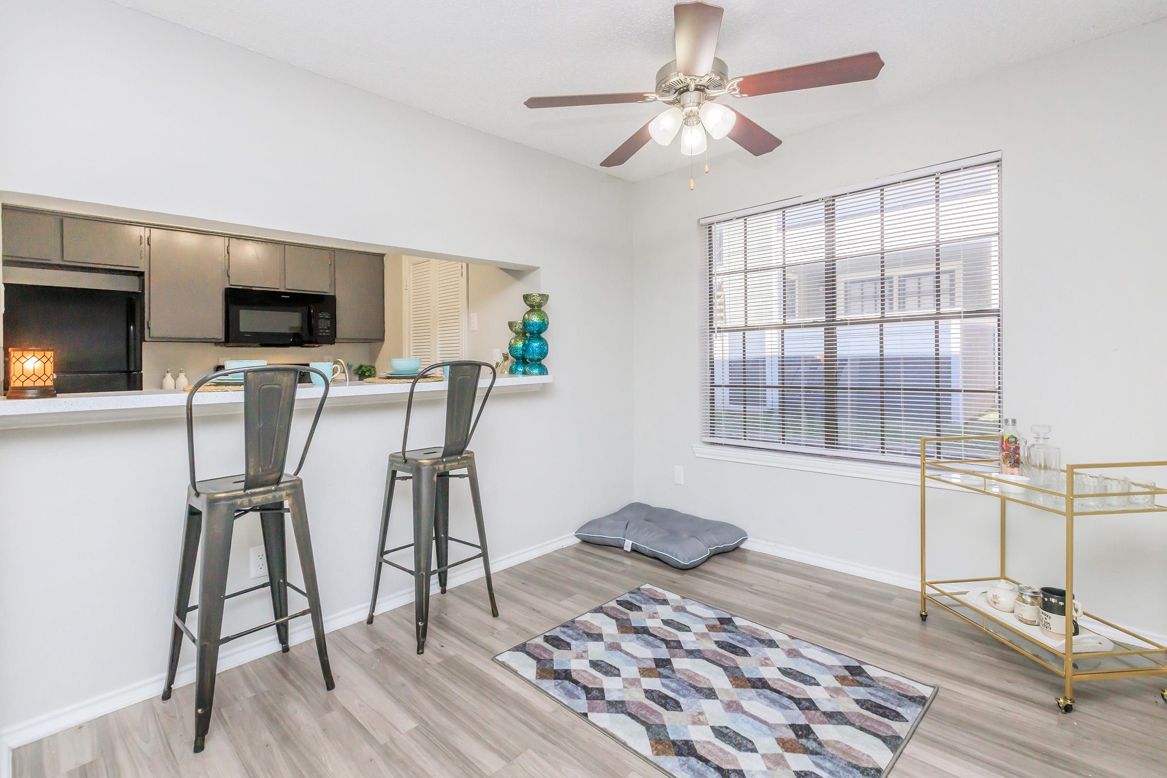 An interior view of a modern kitchen and dining area featuring two bar stools at a counter, a decorative rug on the floor, a window with blinds, and a stylish side table with decorative items. The space has neutral walls and wood-look flooring, illuminated by a ceiling fan.