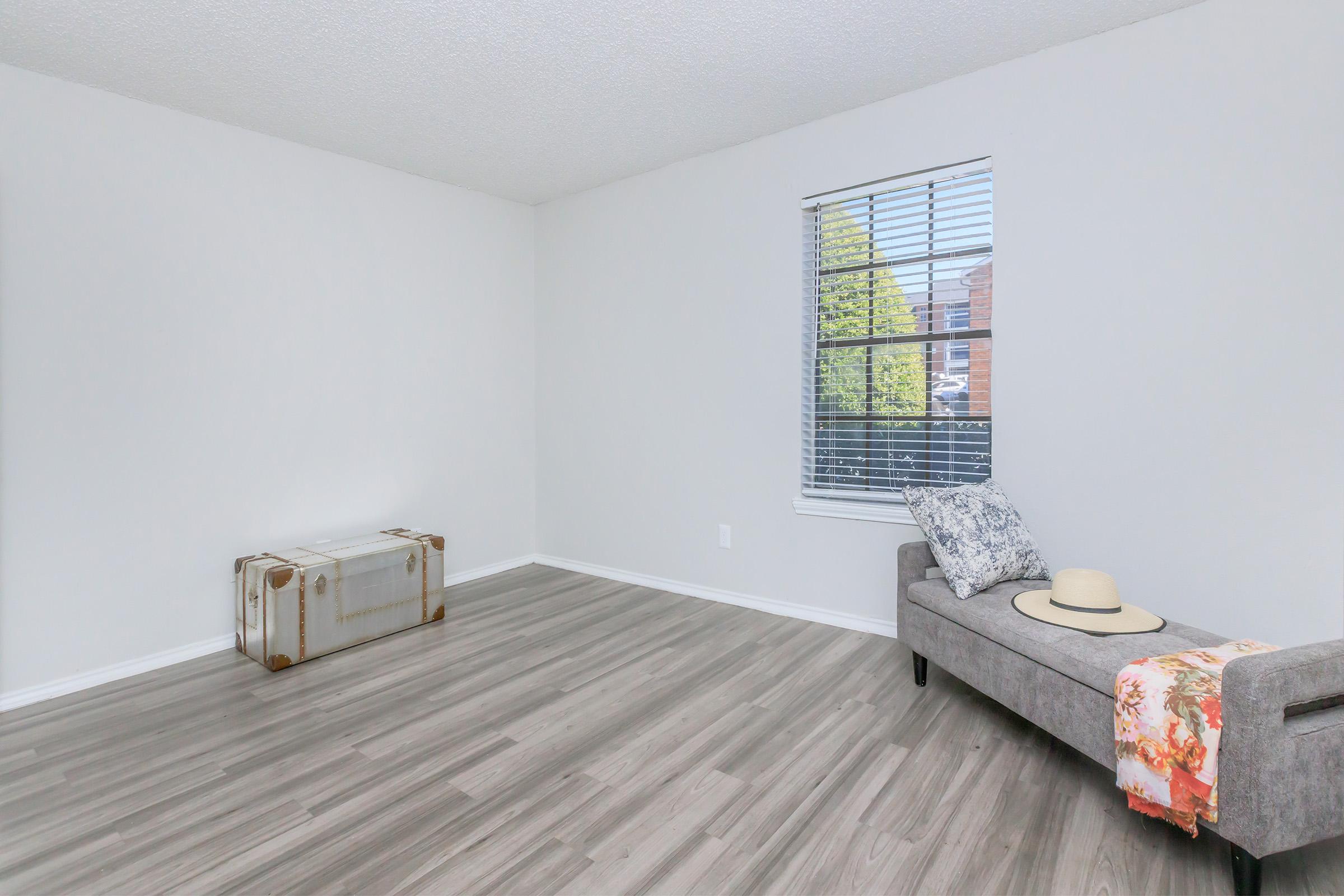 A spacious, empty room featuring light gray walls and a large window with blinds. There is a stylish gray couch with a decorative pillow and a sun hat on it. Next to the couch, there is a vintage suitcase, adding a touch of character to the minimalist space. The flooring is light-colored laminate.