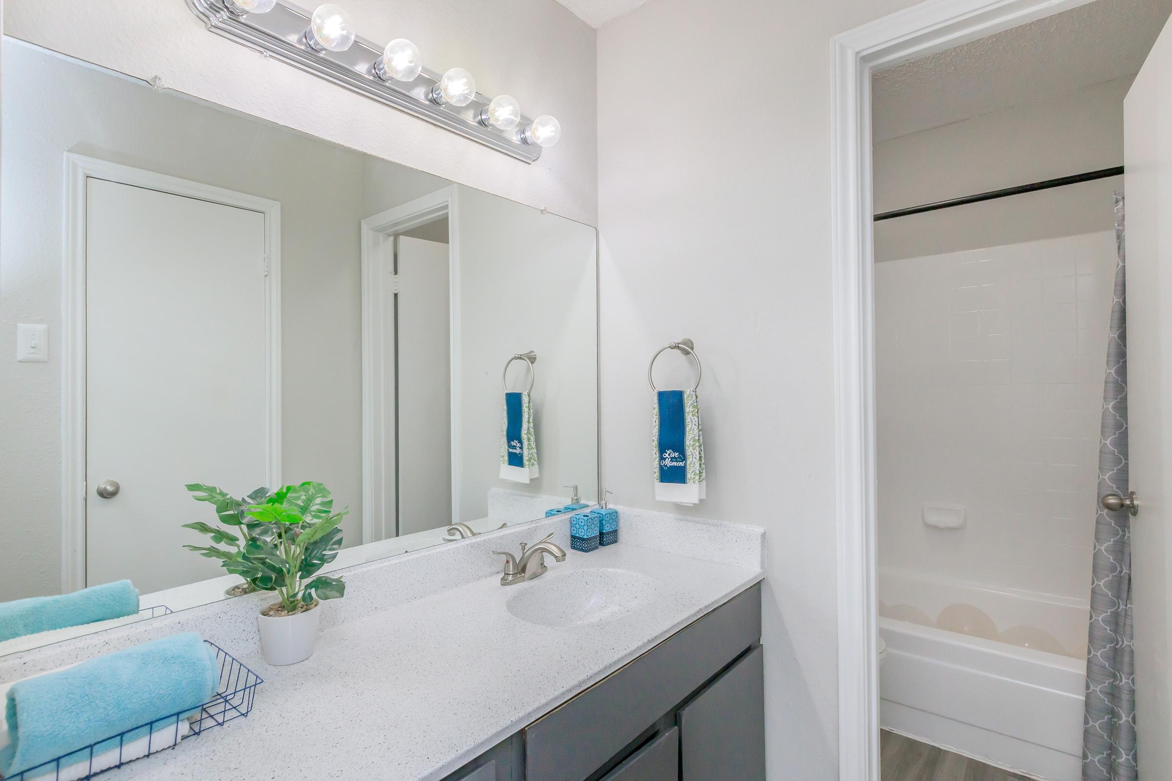 A modern bathroom featuring a double vanity with a large mirror, a stylish light fixture, and a white countertop. There are neatly arranged towels and a small potted plant on the counter. A shower curtain indicates a bathtub/shower area is on the right, and a door leads to another room. The decor is clean and minimalistic.