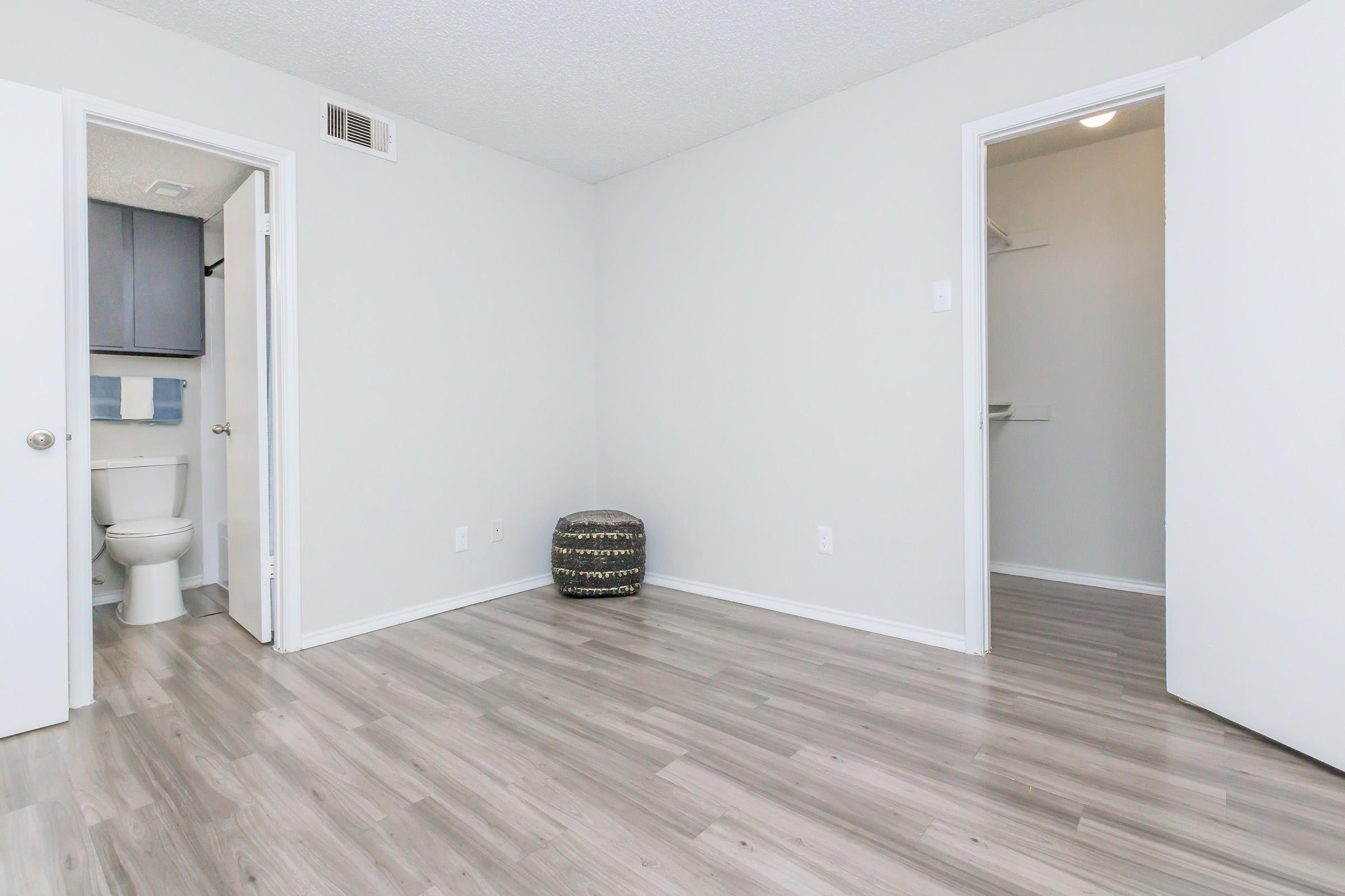 A clean, minimalist room with light gray walls and wood-like flooring. Two doors are visible: one leading to a bathroom with a toilet and the other to a closet. A small decorative stool is placed in the corner, adding a subtle touch to the empty space. Natural light brightens the room.