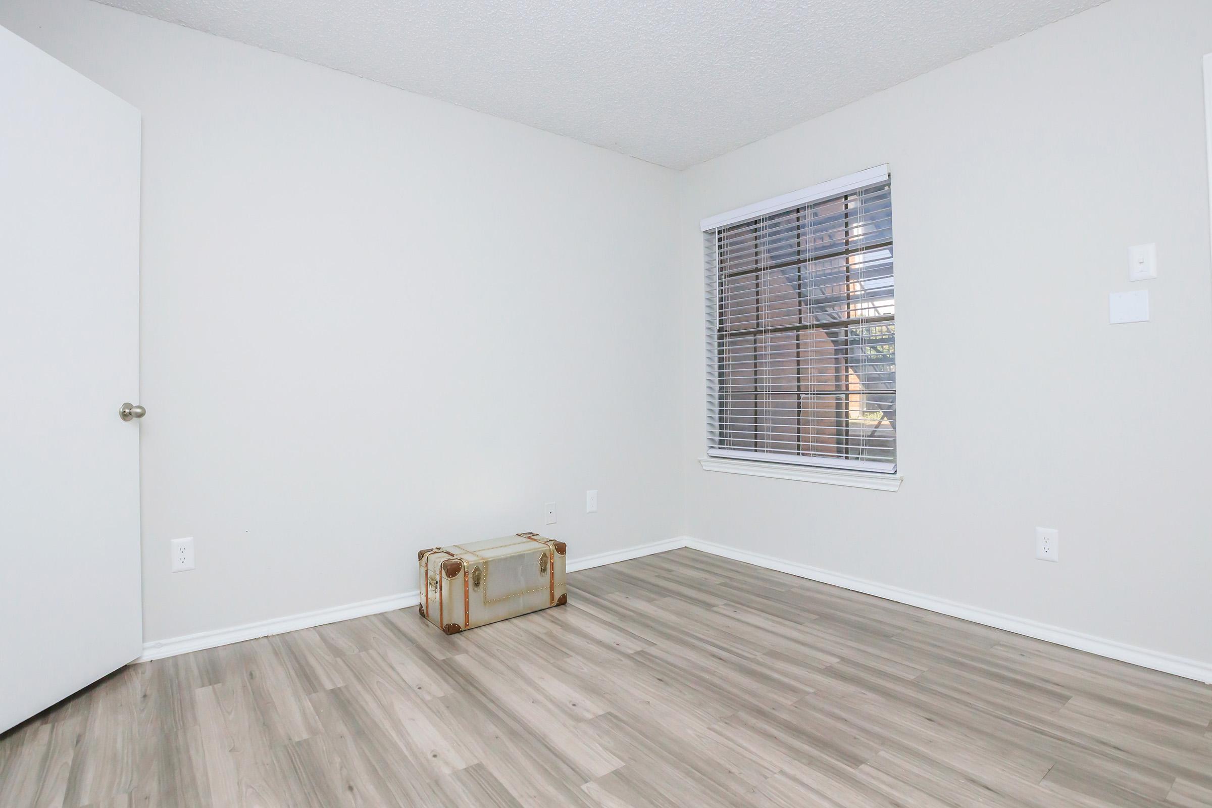 A spacious, empty room with light-colored walls and laminate flooring. There's a closed white door on the left and a window with blinds allowing natural light in. A vintage suitcase is placed on the floor, adding a touch of character to the otherwise minimalistic space.