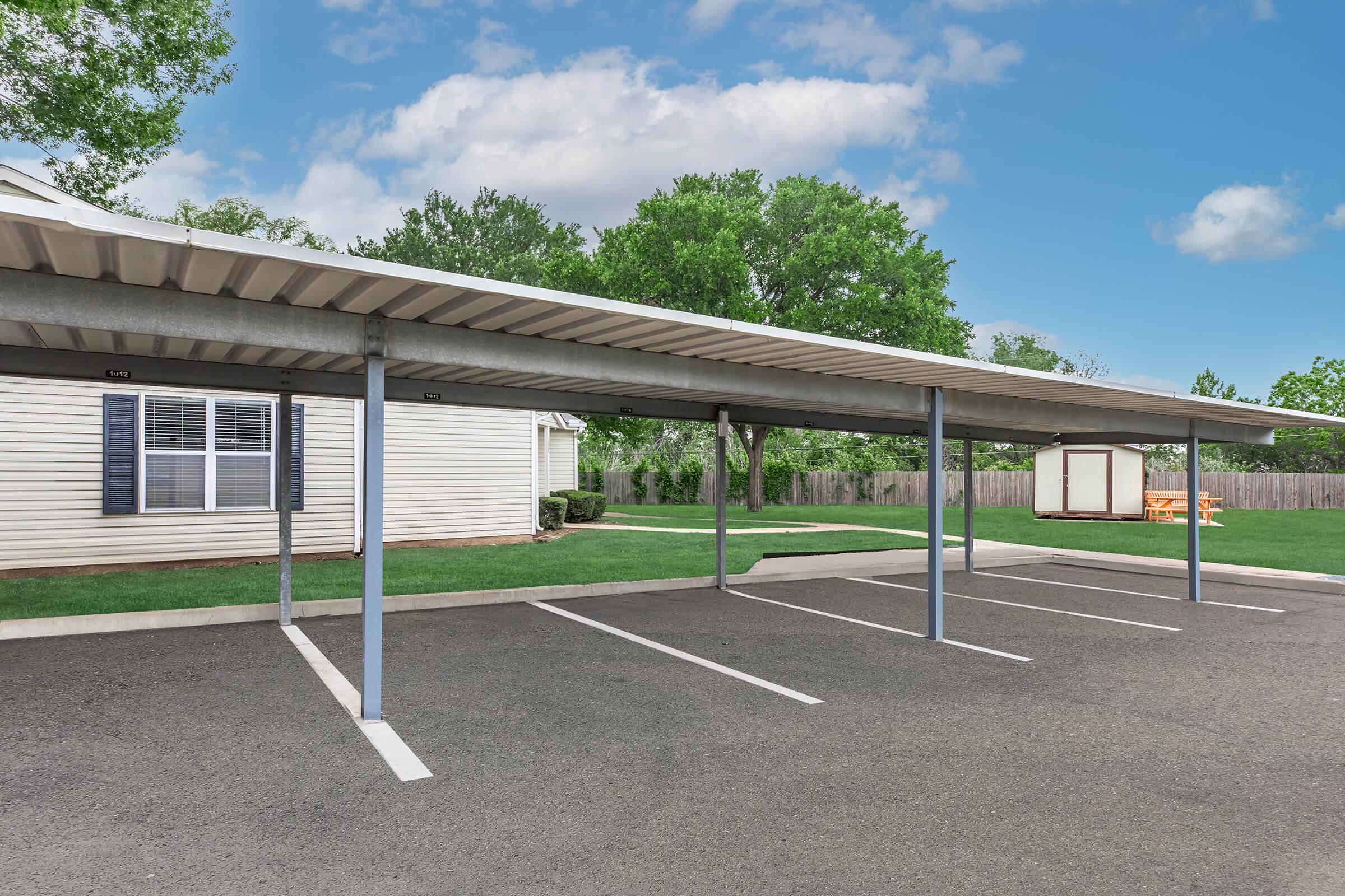 A covered parking area with multiple spaces, featuring metal canopies supported by vertical beams. In the background, there is a single-story building with windows and a grassy area. A wooden structure is visible further back, surrounded by trees and a fence under a partly cloudy sky.