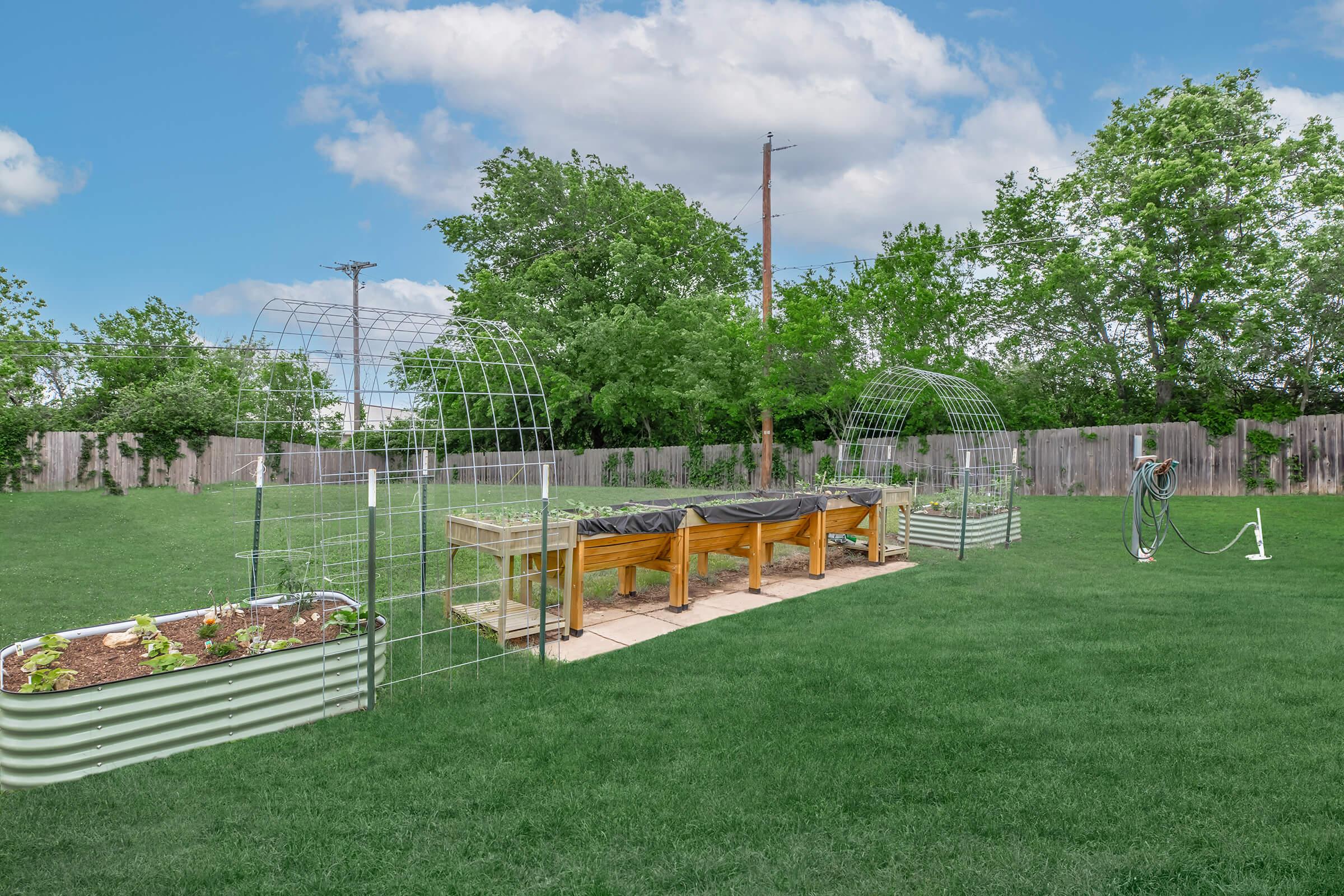 A green backyard featuring two raised garden beds with trellises, lined with pavers. The beds are planted with various seedlings. A hose for watering is visible, and lush trees provide background greenery under a cloudy blue sky. The area is open, with a wooden fence enclosing the space.