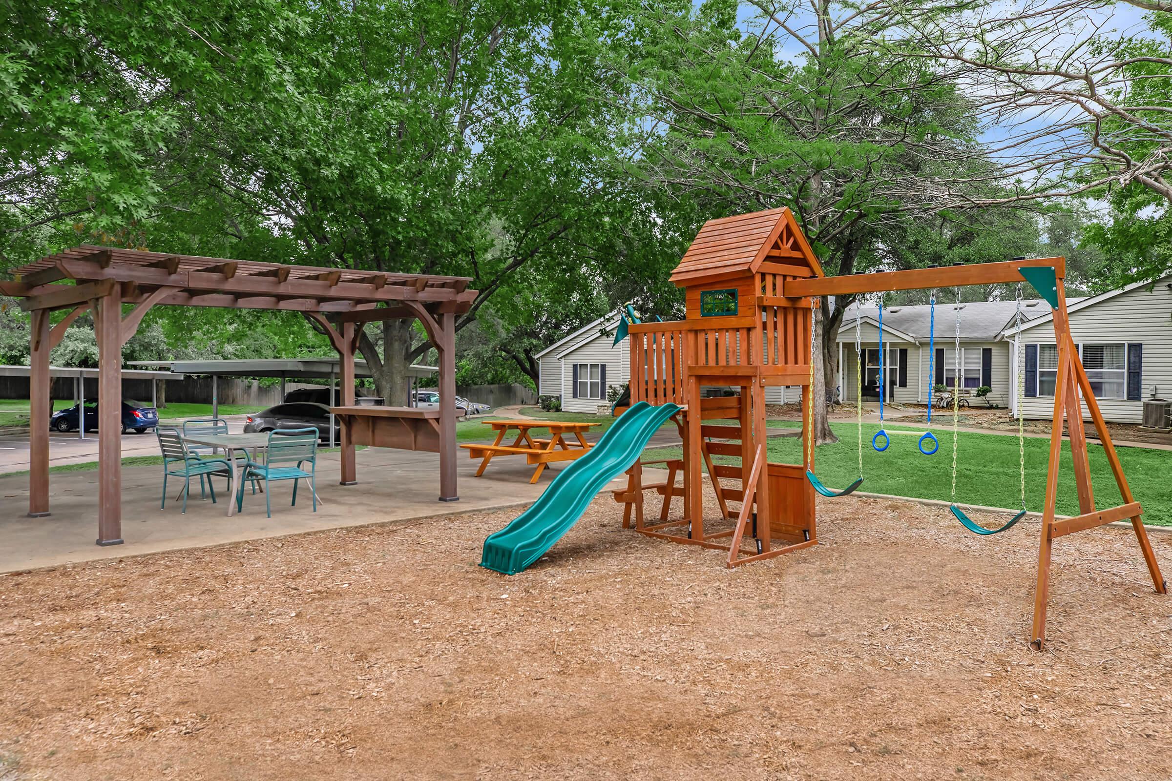 A playground featuring a wooden playset with a slide, swings, and colorful structures. Nearby, there is a shaded picnic area with tables and chairs. The setting includes well-maintained grass and trees, creating a family-friendly outdoor environment. Residential homes are visible in the background.