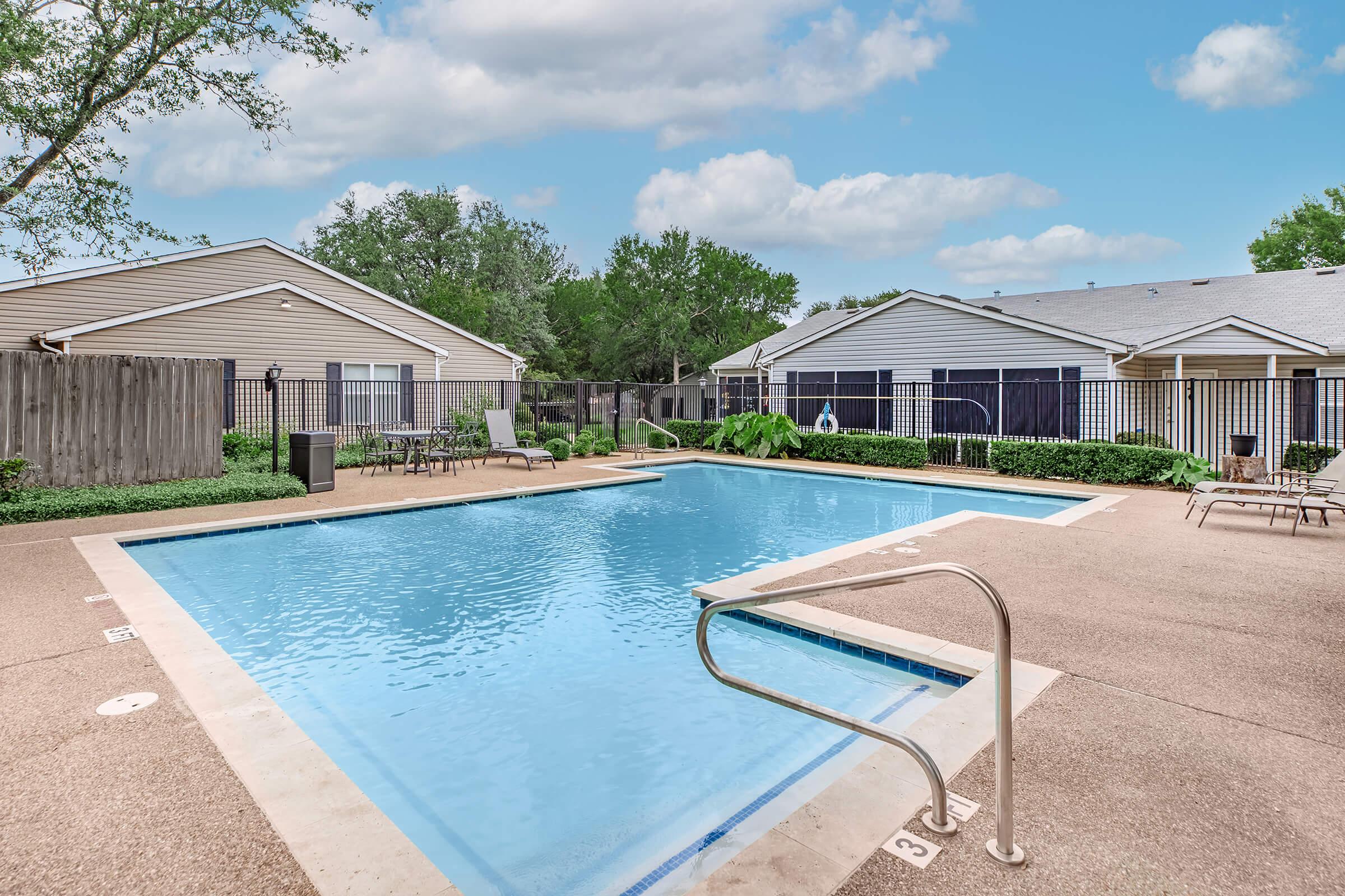 A clear swimming pool surrounded by concrete, with lounge chairs and greenery. Residential buildings are visible in the background, along with a fence for privacy. The sky is partly cloudy, creating a serene outdoor atmosphere.