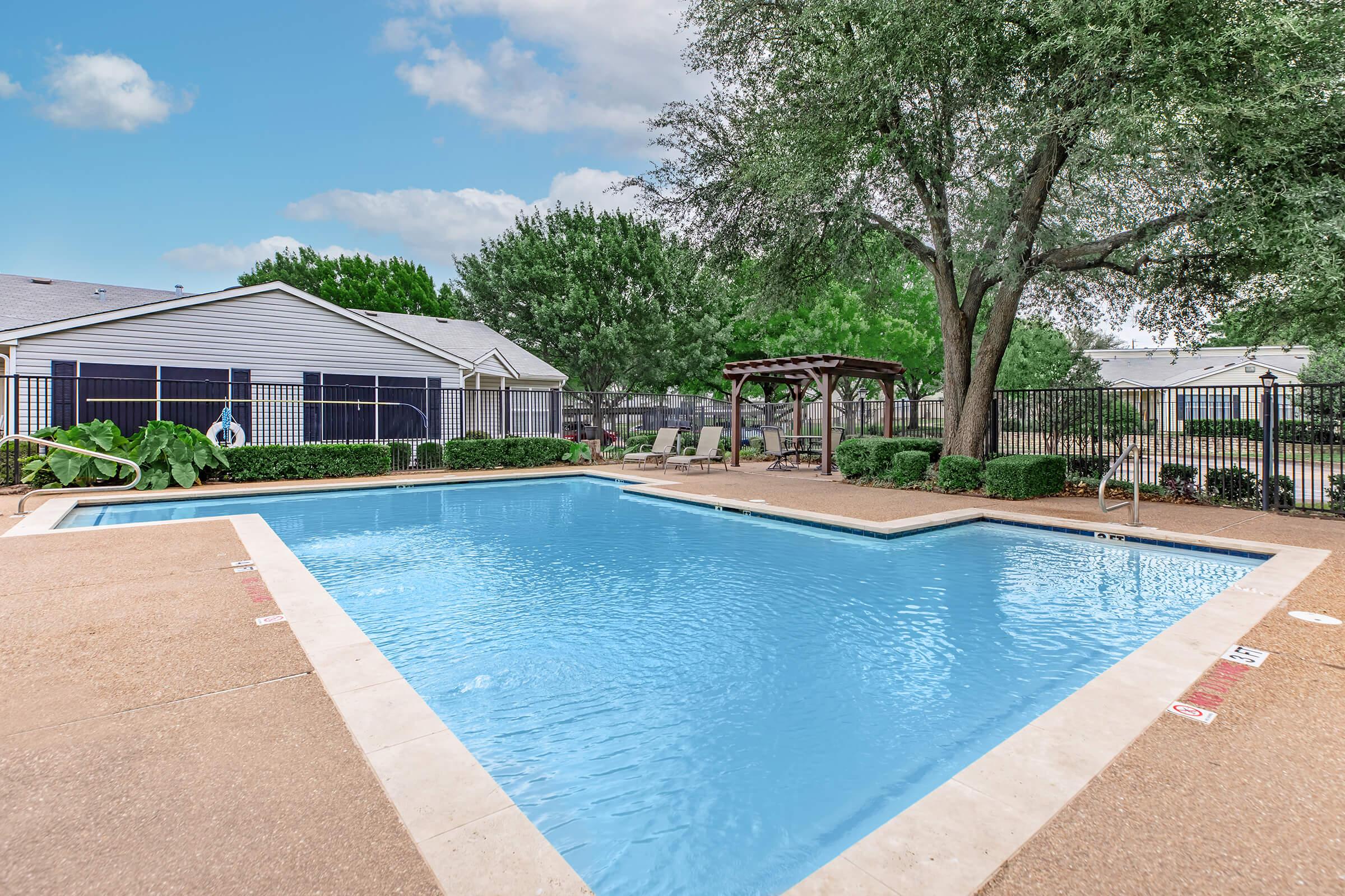 A serene outdoor swimming pool surrounded by landscaped greenery. The clear blue water reflects the sky, with a gentle slope leading into the pool. Notable features include a pergola and lounge chairs nearby, set against a backdrop of houses and trees in a well-maintained residential area.