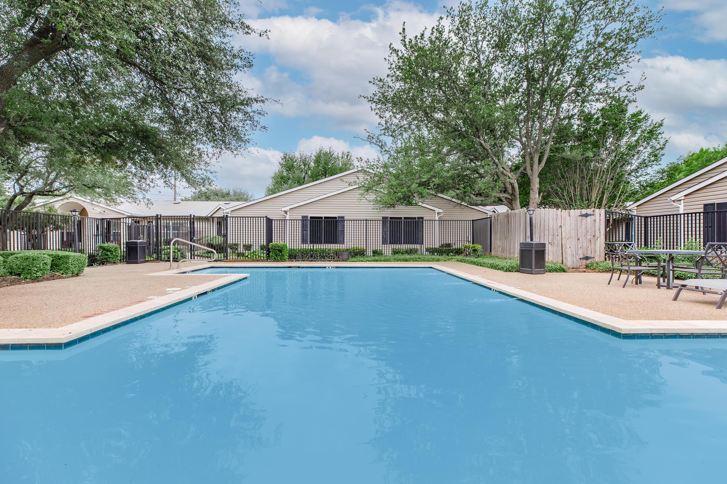 A clear blue swimming pool surrounded by a well-maintained area featuring green shrubs, trees, and a fence. There are lounge chairs and tables nearby, and quaint, single-story residential buildings in the background under a partly cloudy sky.