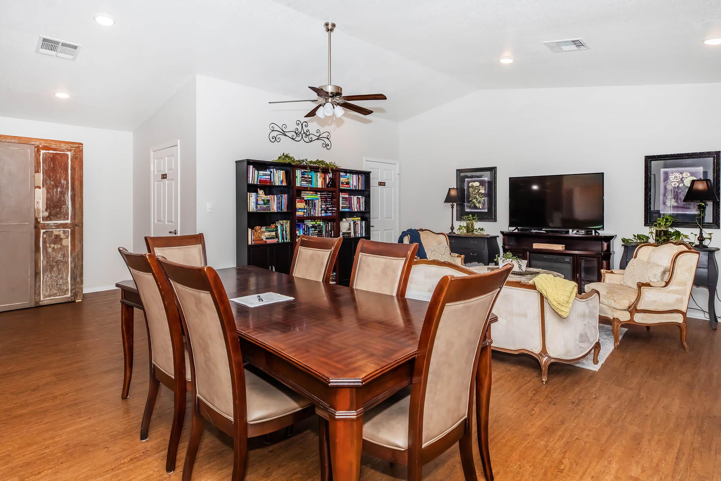 A spacious dining area featuring a large wooden table surrounded by elegant chairs. In the background, there is a cozy living room with a TV and a bookshelf filled with books. The room has light-colored walls and wooden flooring, creating a warm and inviting atmosphere.