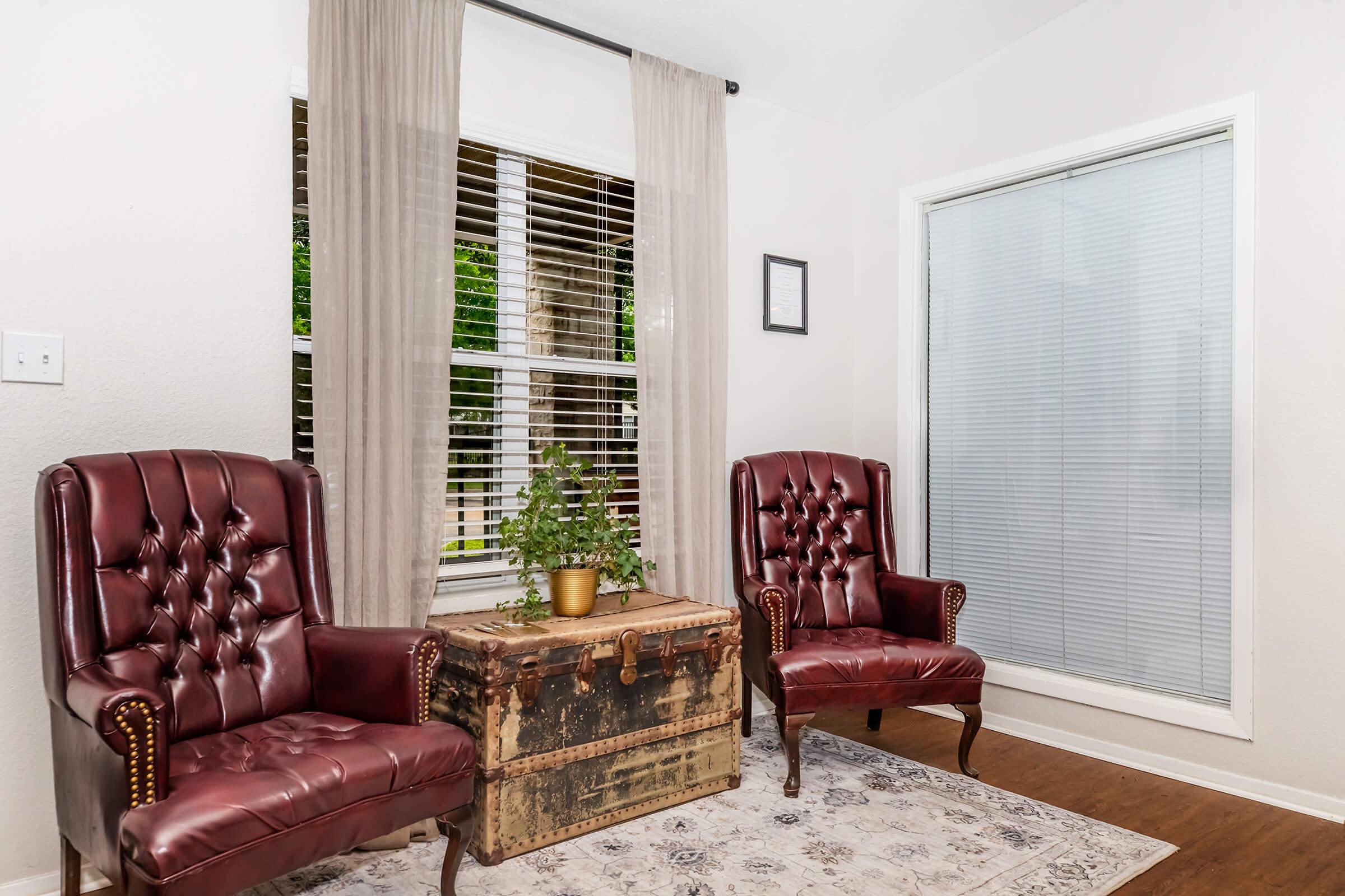 Two burgundy tufted armchairs flanking a vintage wooden trunk in a cozy room. A potted plant sits on the trunk, and large windows with sheer curtains allow natural light to brighten the space. The floor is wooden, and there is a patterned area rug underneath the trunk.