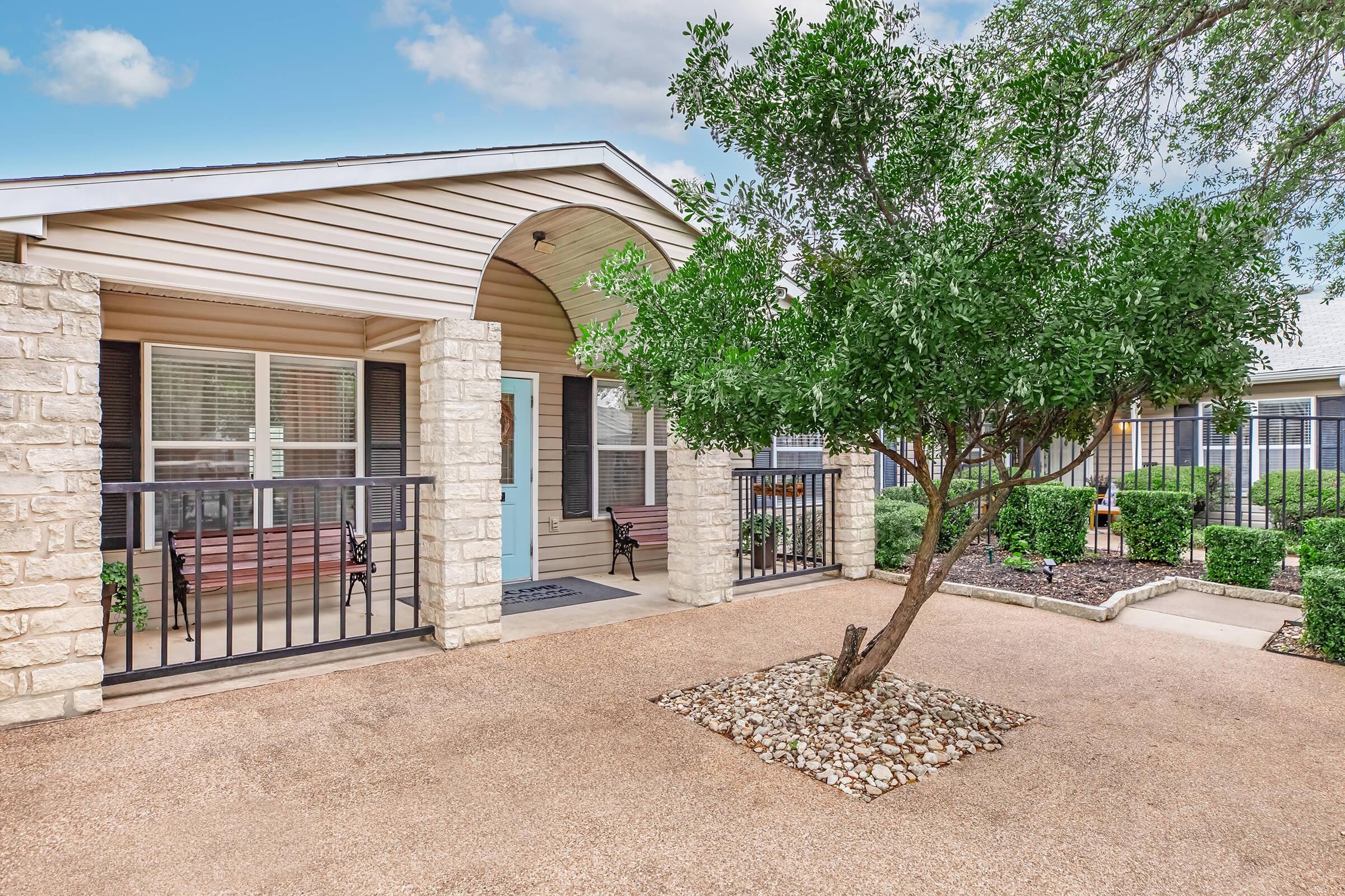 A charming entrance to a home featuring a covered porch with seating, a well-maintained garden, and a small decorative tree. The pathway is lined with gravel, and there are manicured shrubs and stone arrangements enhancing the outdoor space. The overall aesthetic is welcoming and inviting.