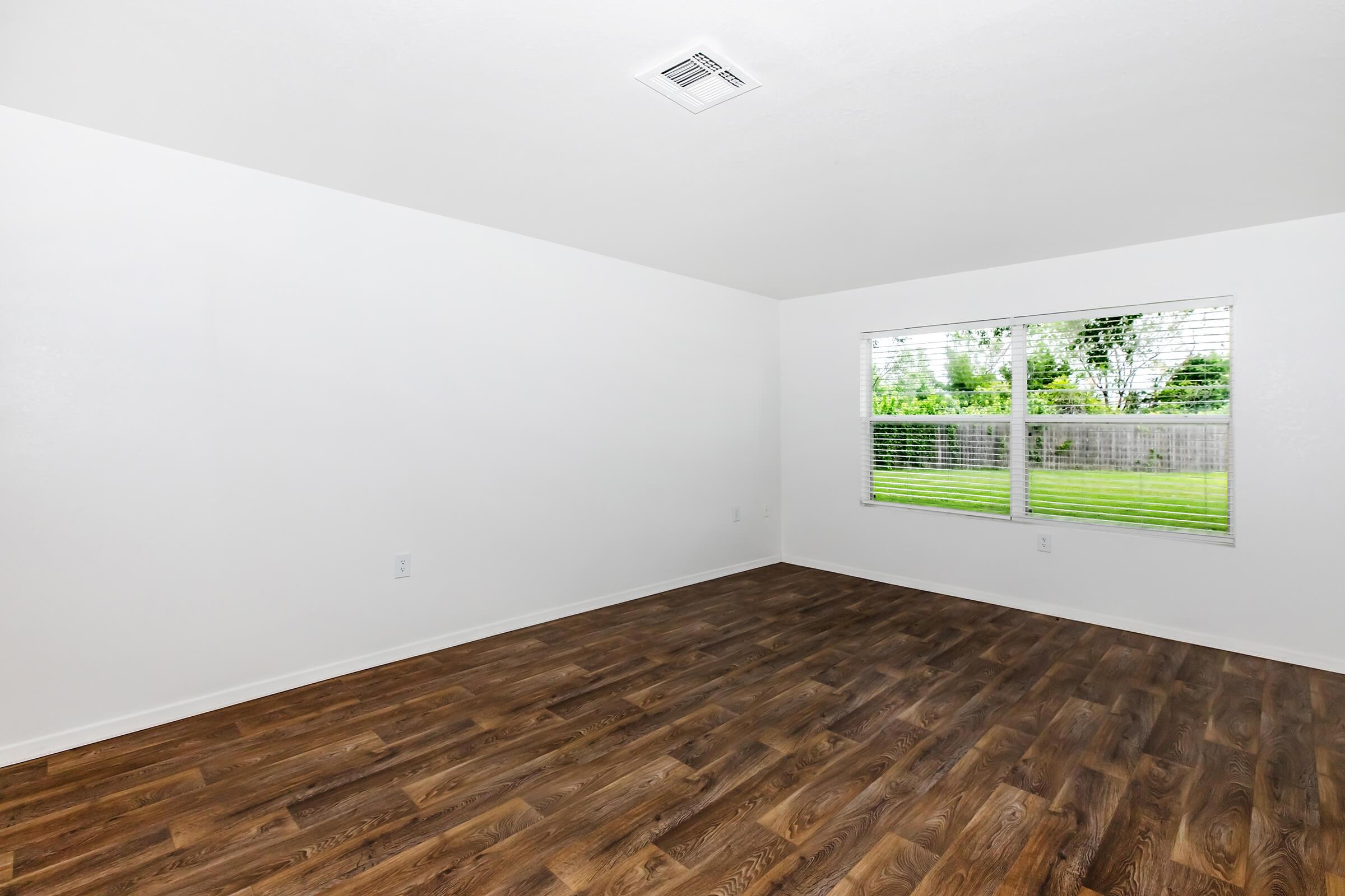 Empty room with light-colored walls and a window featuring white blinds. The flooring is a dark wood laminate, and the view outside the window shows greenery. The space is bright and uncluttered, creating a fresh and airy atmosphere.