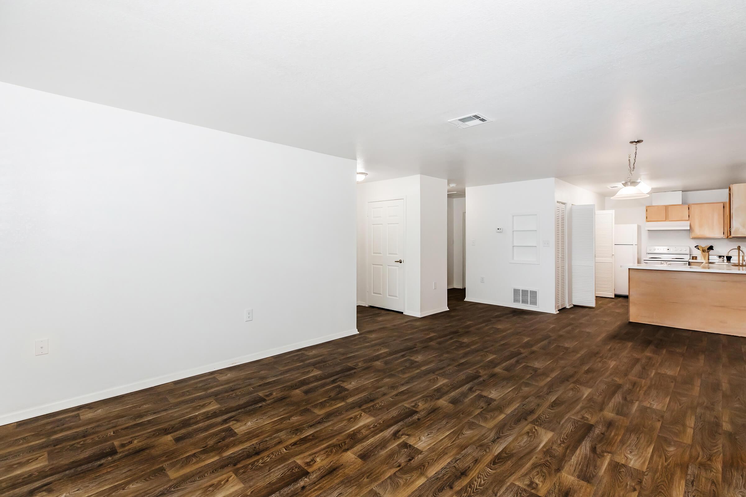 An empty apartment interior featuring a spacious room with light-colored walls and dark wooden flooring. The kitchen is visible in the background, equipped with light wood cabinets and appliances. Natural light brightens the space, creating a warm and inviting atmosphere.