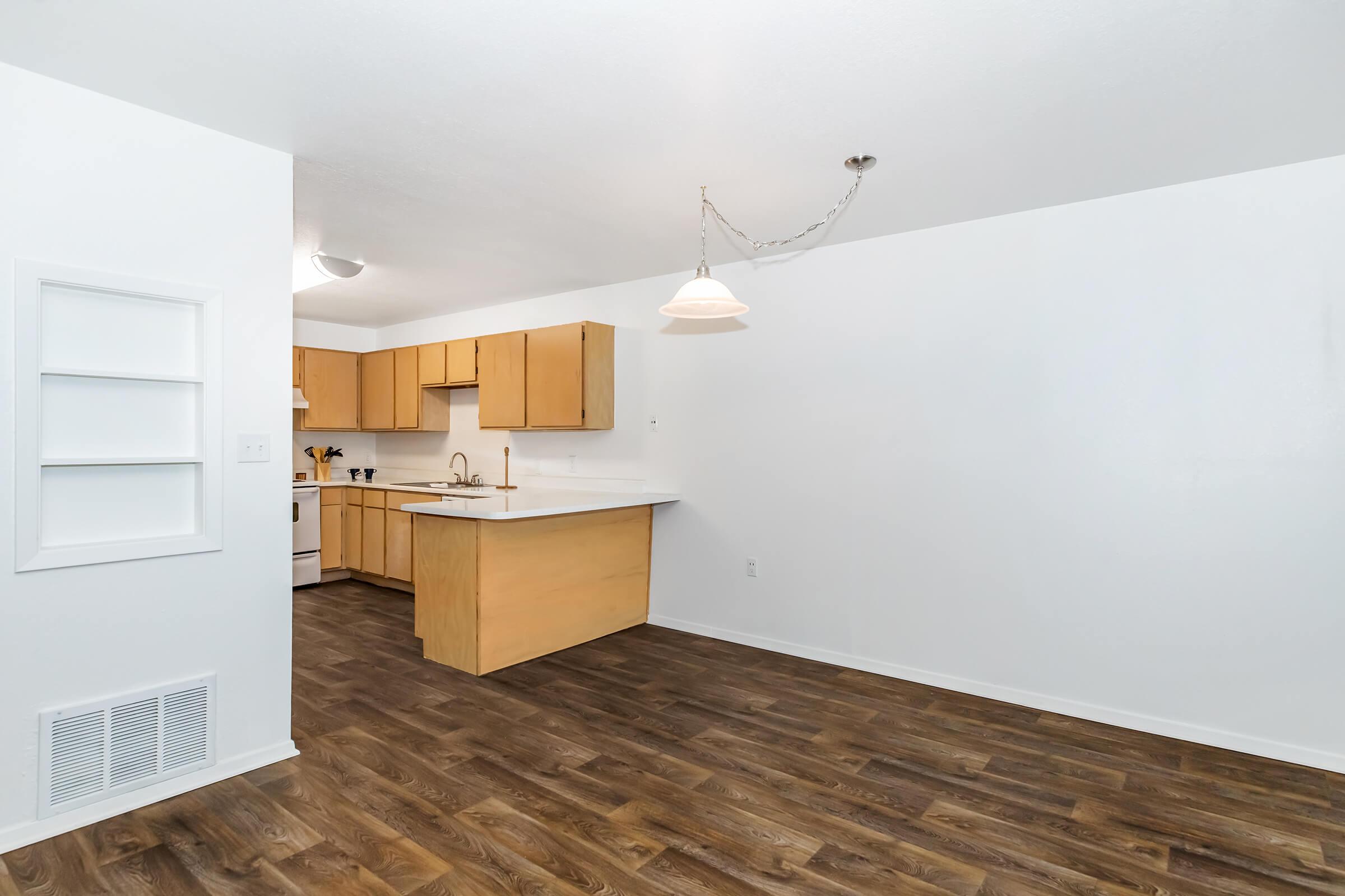 Bright and spacious kitchen area with light wooden cabinets and a white countertop. The room features a pendant light hanging from the ceiling and has modern laminate flooring. There are appliances visible, including a stove and refrigerator, with a small shelf on one wall. The walls are painted white, creating an airy feel.