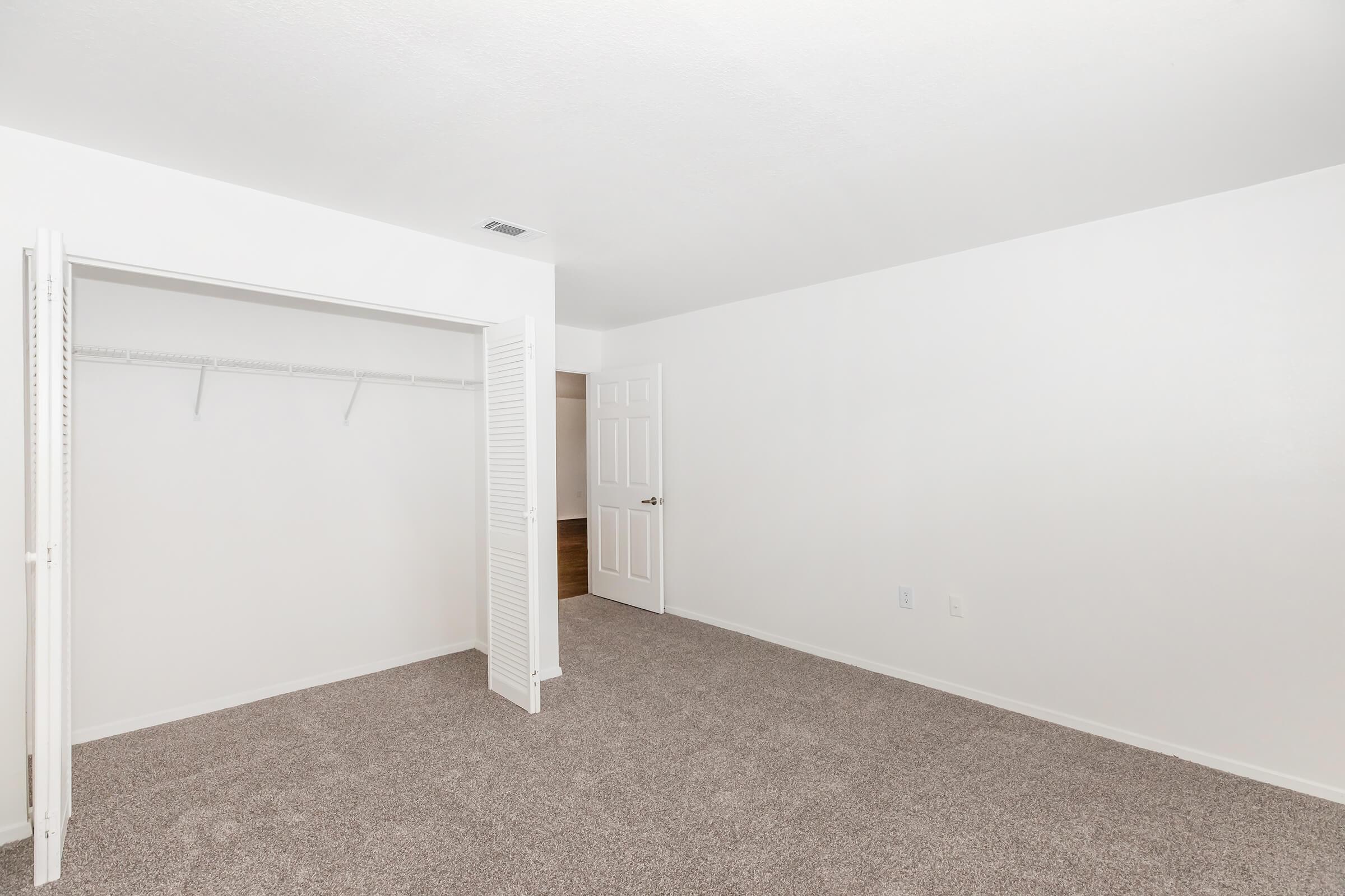 A spacious and empty room with light-colored walls and carpet. It features an open closet with a shelf, a closed door leading to another room, and a bright, well-lit atmosphere. The floor is carpeted, adding to the room's cozy feel.