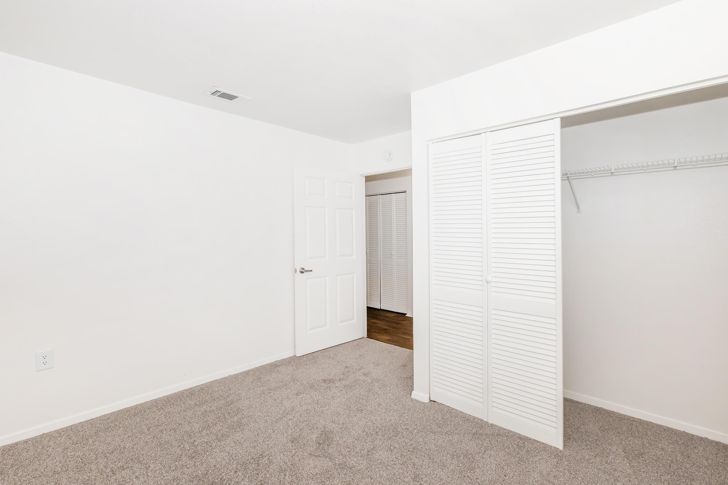 Empty room with light-colored walls and beige carpet. A closed white door is on the left side, while another door with louvered panels is on the right, leading to a closet area. The room has a bright, clean appearance with no furniture or decorations.