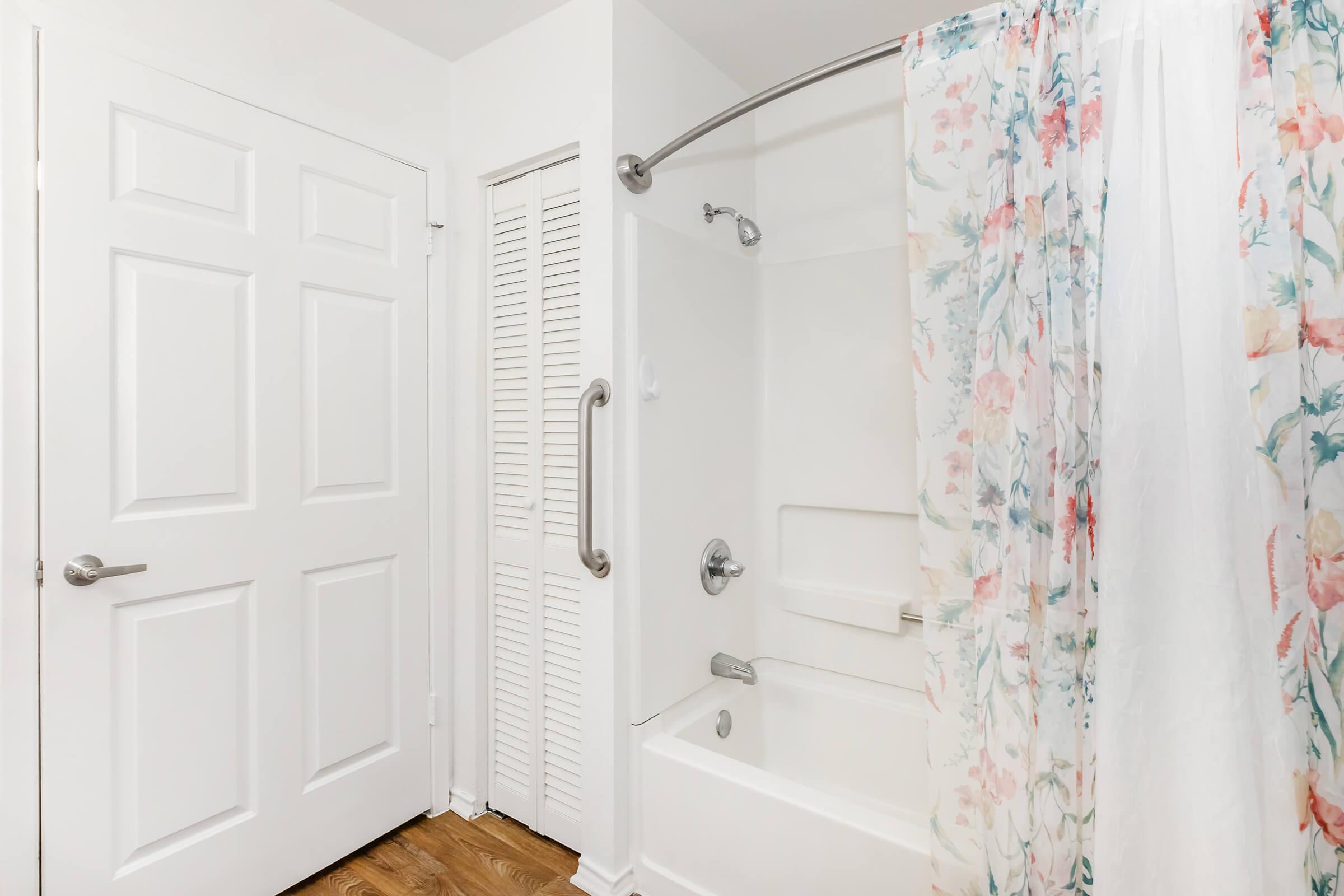 A clean bathroom featuring a white bathtub with a shower curtain adorned with floral patterns. There is a white door leading to another room and a closet with louvered doors. The flooring is wooden, contributing to a light and airy ambiance.