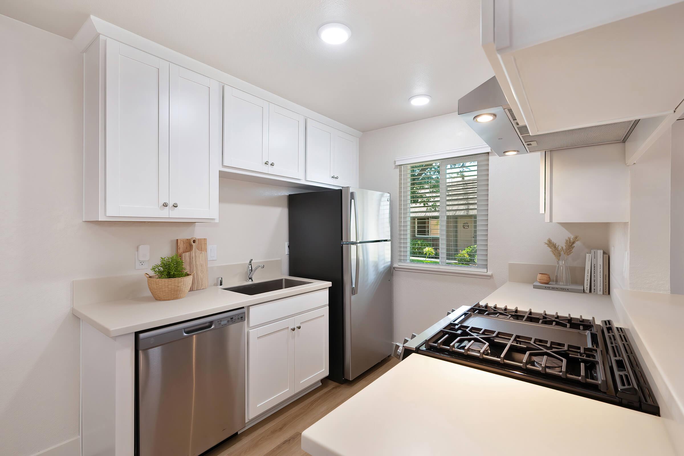Modern kitchen featuring white cabinetry, a stainless steel refrigerator, a sink, and a stove with a hood. The countertop is light-colored, and there's a window allowing natural light and a view of greenery outside. Soft lighting illuminates the space, creating a clean and inviting atmosphere.