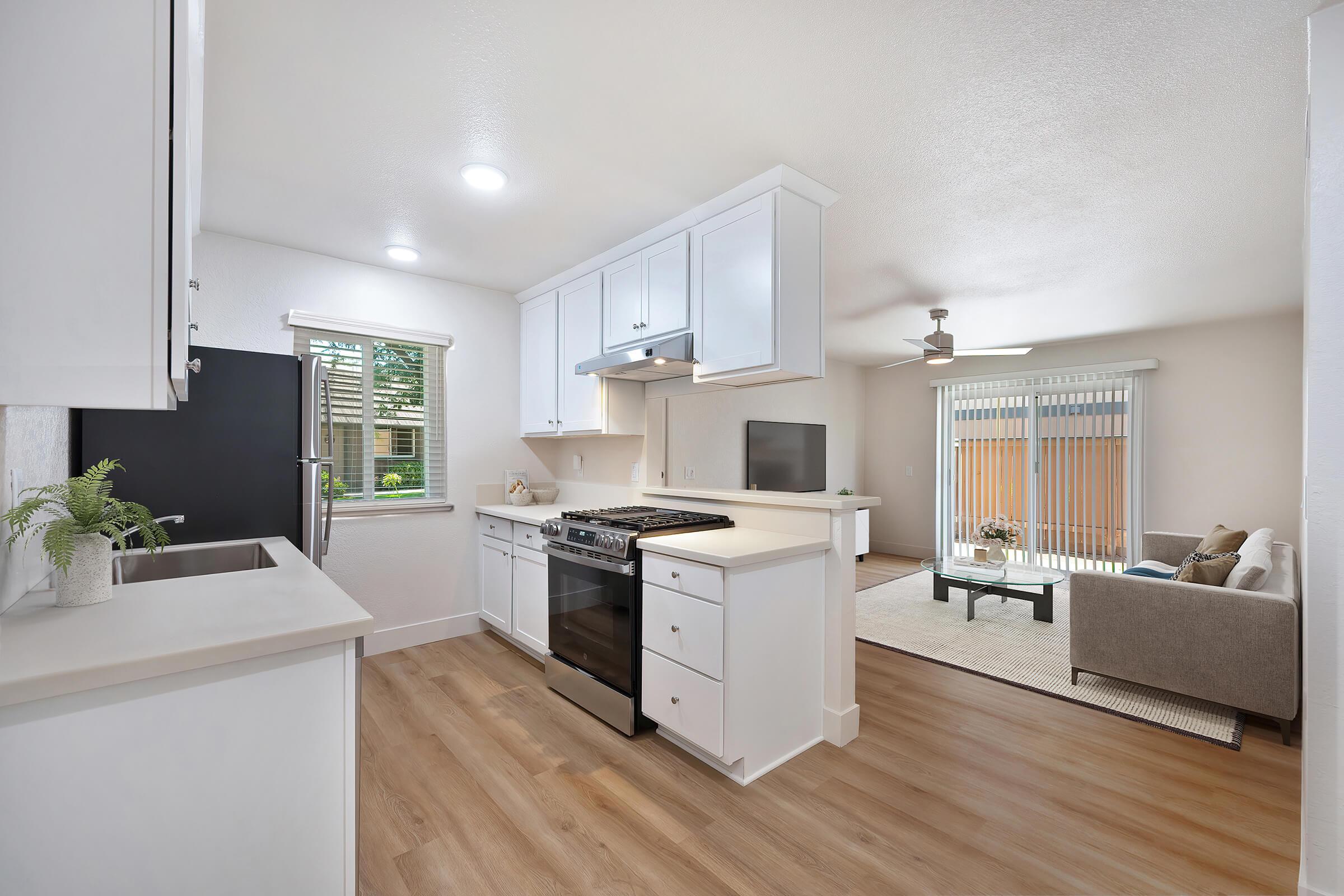 A modern kitchen featuring white cabinets, a gas stove, and a window letting in natural light. The kitchen opens up to a spacious living area with a ceiling fan, a comfortable sofa, and a coffee table, with sliding glass doors leading to an outdoor space. The flooring is light wood, creating a warm ambiance.