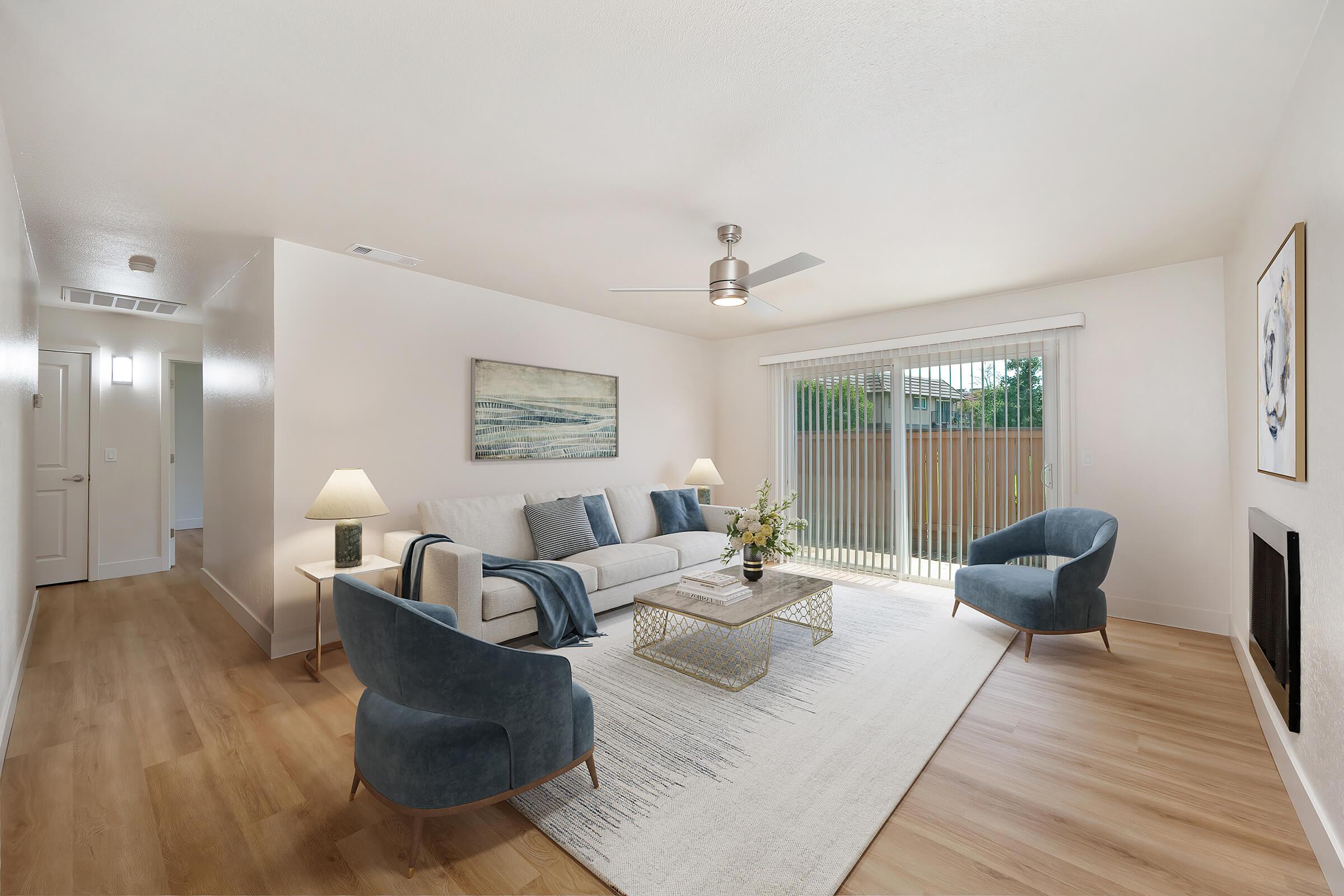 A modern living room featuring a light-colored sofa with blue and gray cushions, two stylish blue chairs, and a decorative coffee table. Natural light streams in through sliding glass doors, revealing a view of greenery outside. Soft, neutral walls and a rug contribute to a cozy atmosphere.