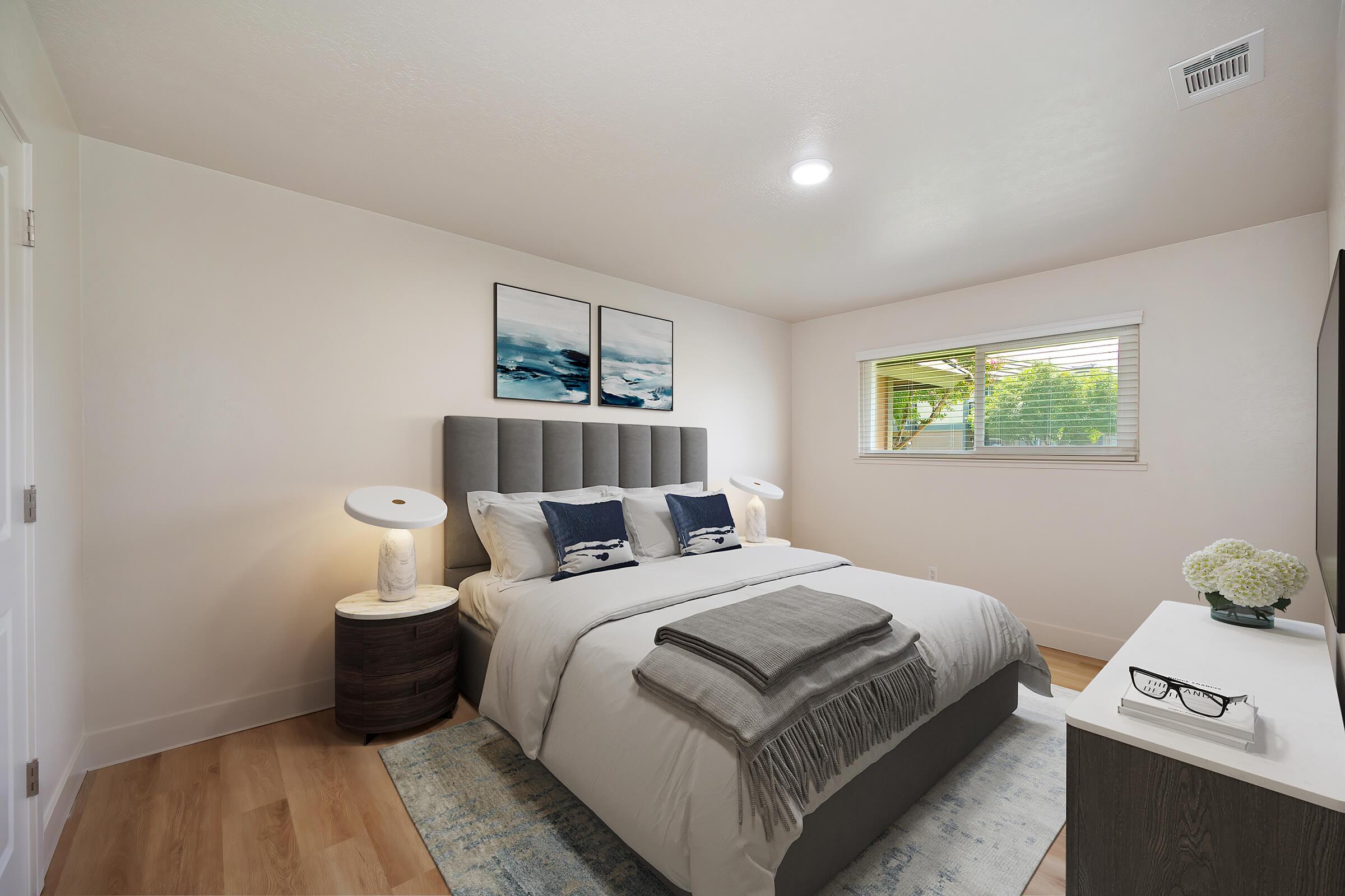 A modern bedroom featuring a queen-sized bed with gray bedding and decorative pillows. There are two bedside lamps on round nightstands, and three framed ocean-themed artworks on the wall. A window allows natural light, and a small dresser with a decorative vase is present, set on light wood flooring.